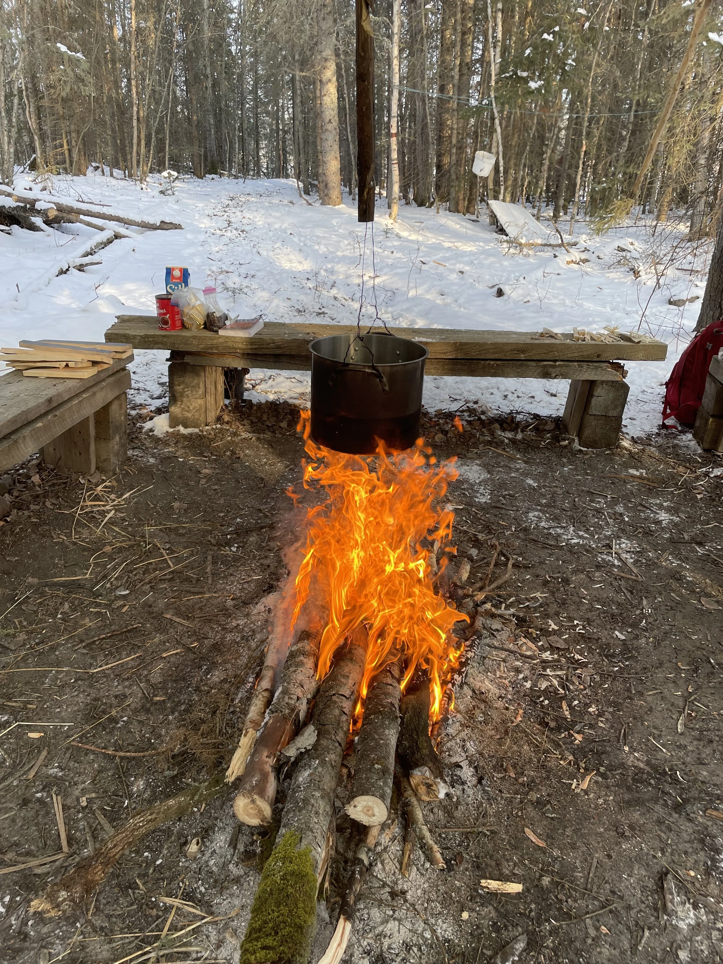 A campfire with a hanging pot over it in a snowy forest setting, with wooden benches and supplies nearby.