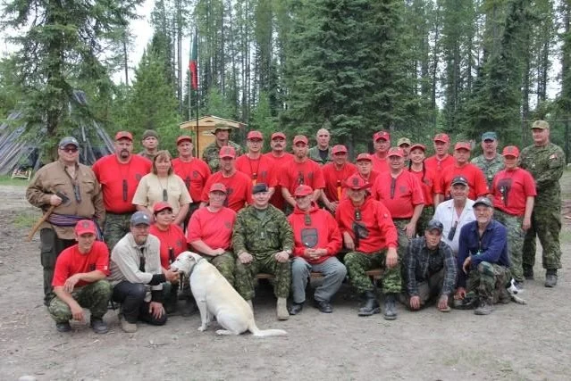 A course photograph of Canadian Rangers and instructors sitting with Mors Kochanski outdoors in a forest, with a dog, some seated and others standing.
