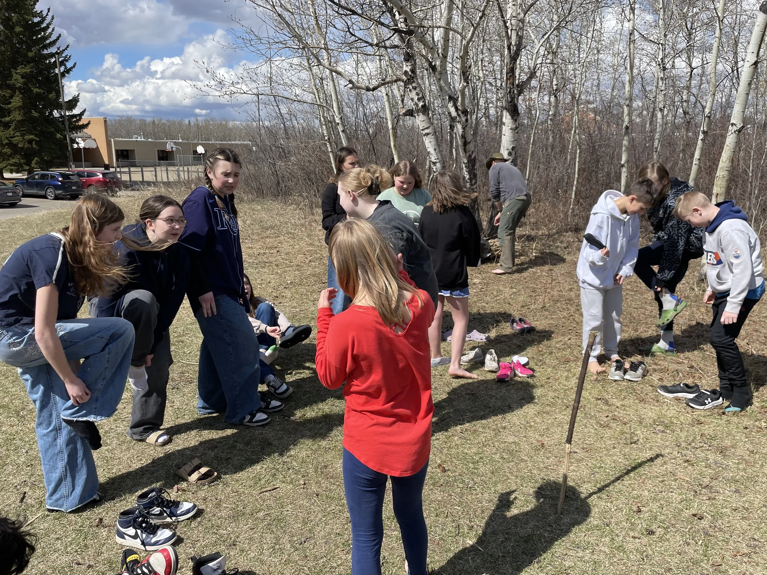 Group of young people outdoors, some barefoot, putting on shoes on a grassy area near trees on a partly cloudy day.