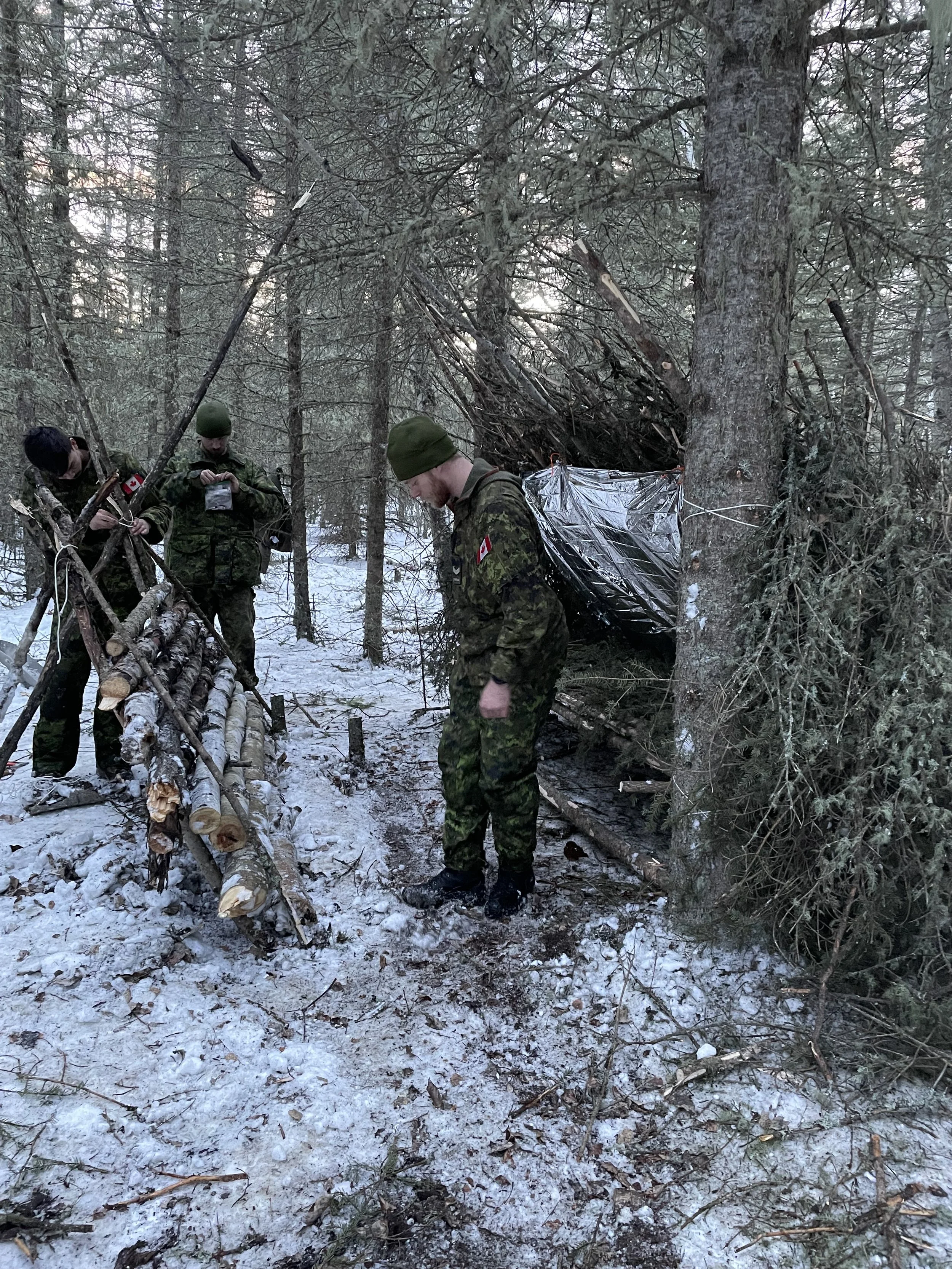 Three soldiers in camouflage uniforms and green hats in a snow-covered forest, inspecting a makeshift shelter built with wood and a tarp.