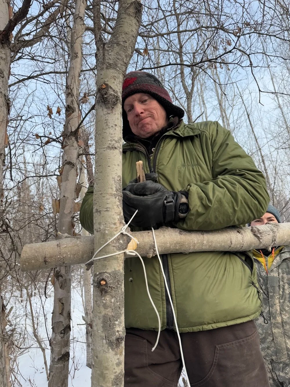 An instructor in a green jacket and knit cap standing outdoors in a winter forest, helps to make an improvised shelter.