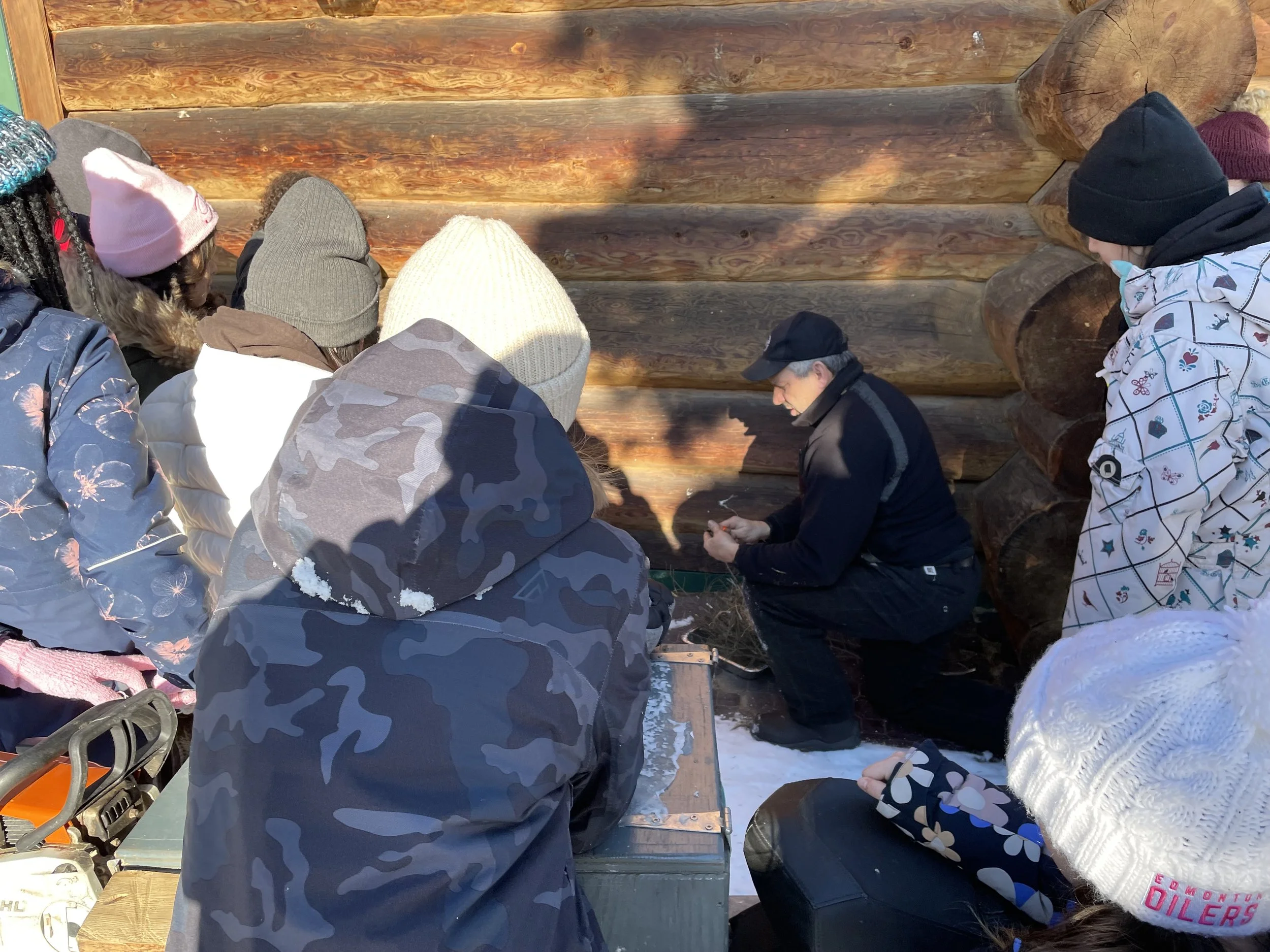 A group of children in winter clothing gathered around a man kneeling in front of a wooden log wall, engaged in an outdoor educational activity.