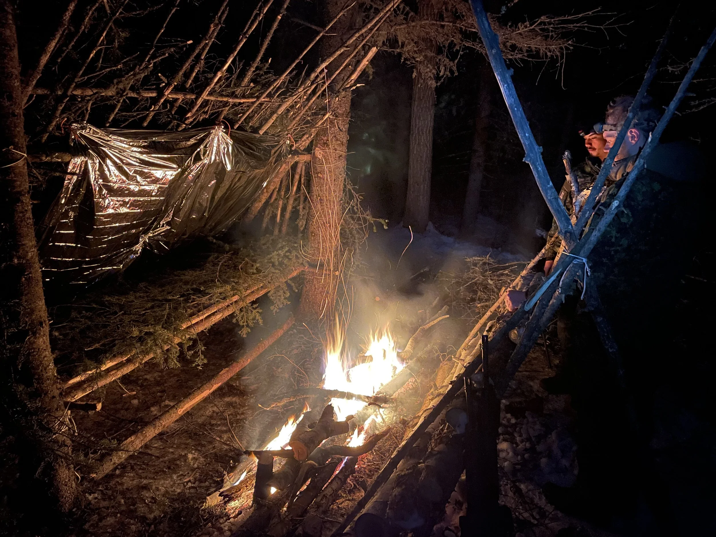 Two individuals outdoors at night around a small campfire in a wooded area, with one person sitting on a wooden structure, possibly a ladder, wearing a headlamp.