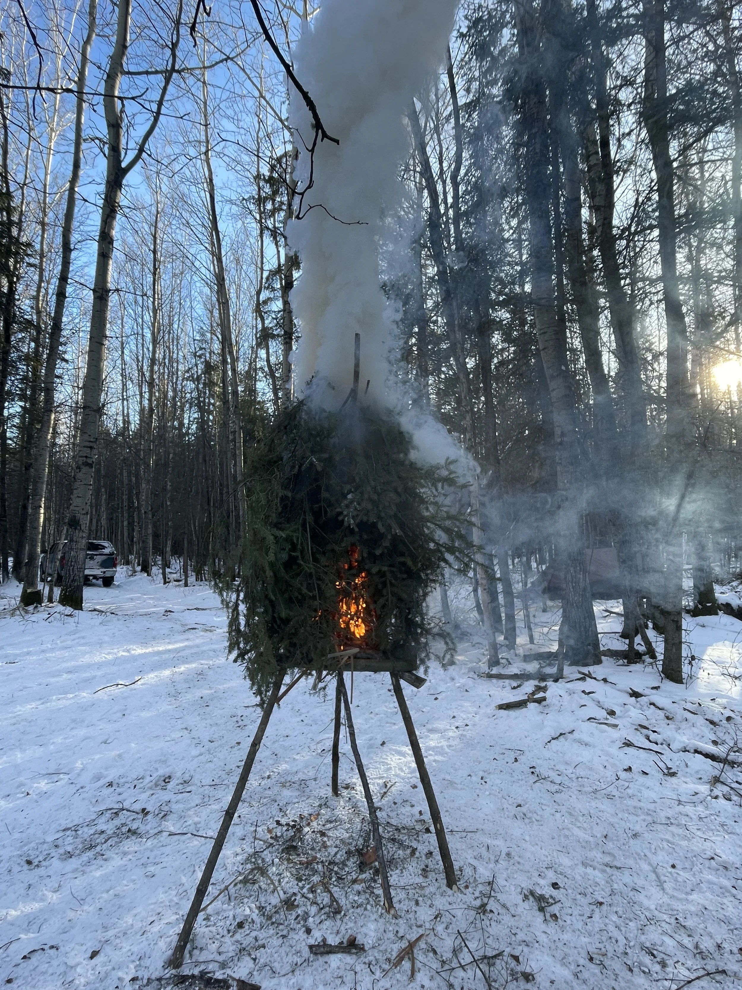 A small fire burning on a tripod in a snowy forest during daylight with smoke rising, surrounded by leafless trees.