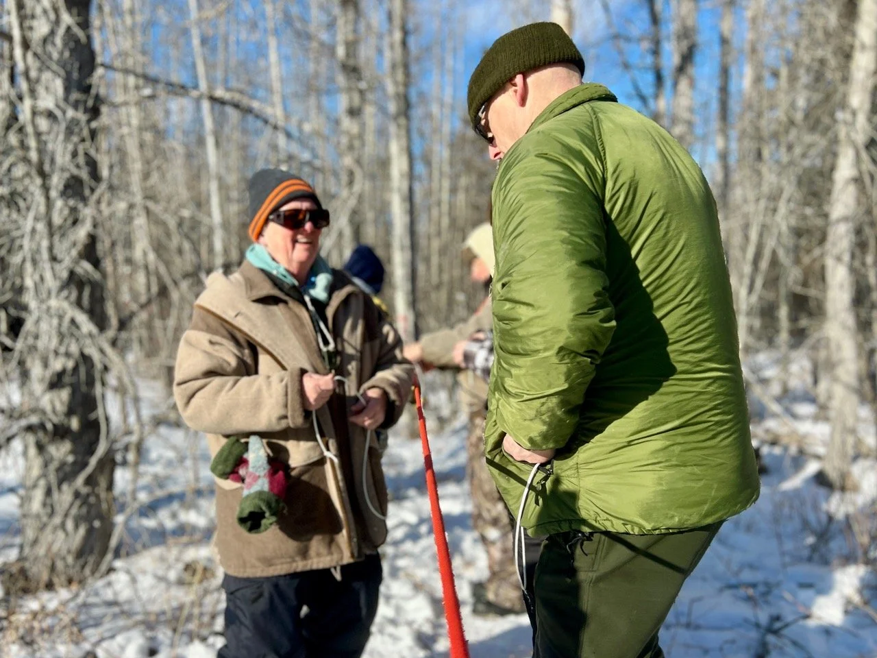 People learning knots in a snowy forest, wearing warm clothing and sunglasses.