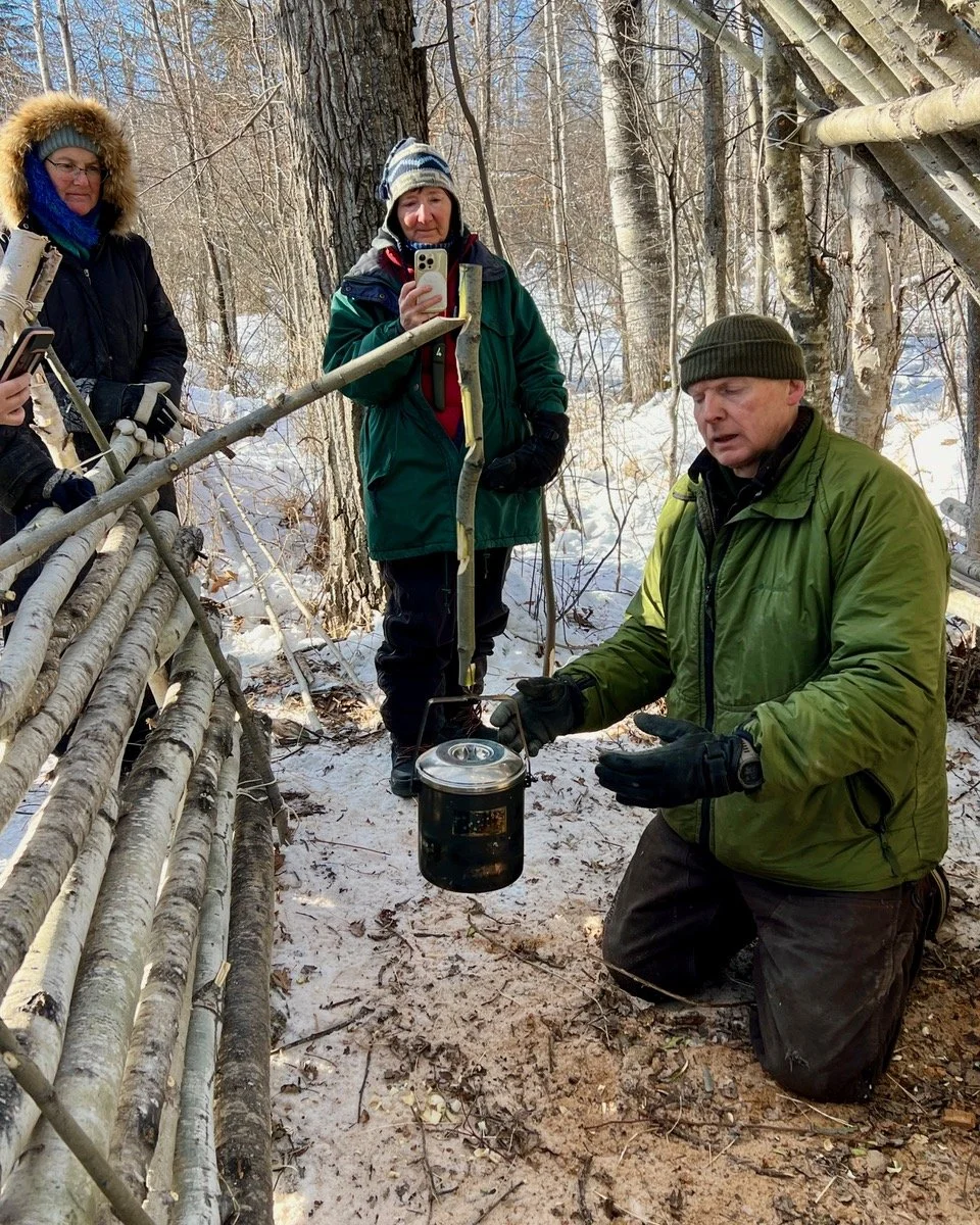 Group of people outdoors in a snowy forest, with the instructor kneeling and holding a metal container, while others observe and take photos.