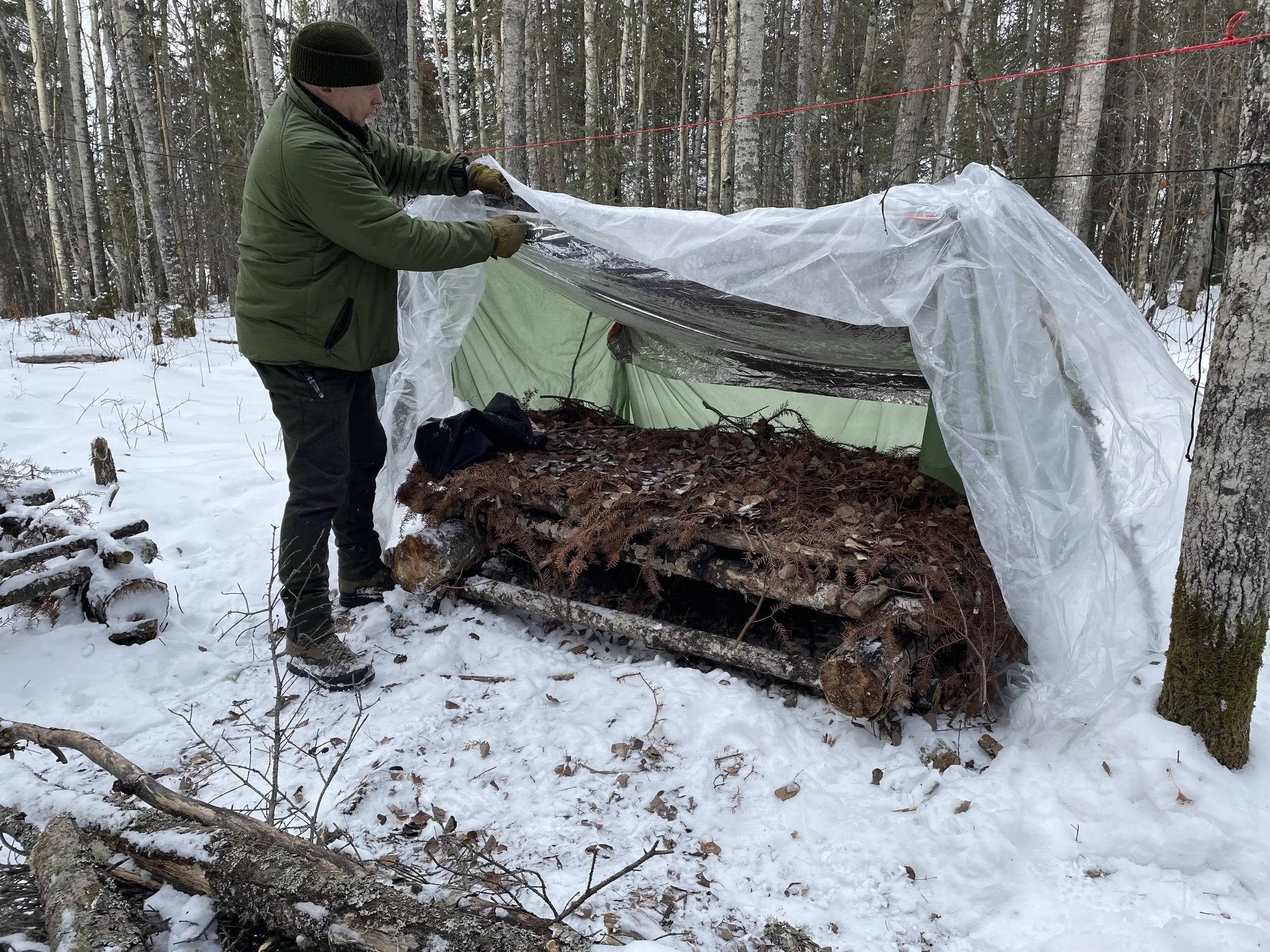 Person setting up a small tarp shelter in a snowy forest, surrounding logs covered with ferns serve as a base.