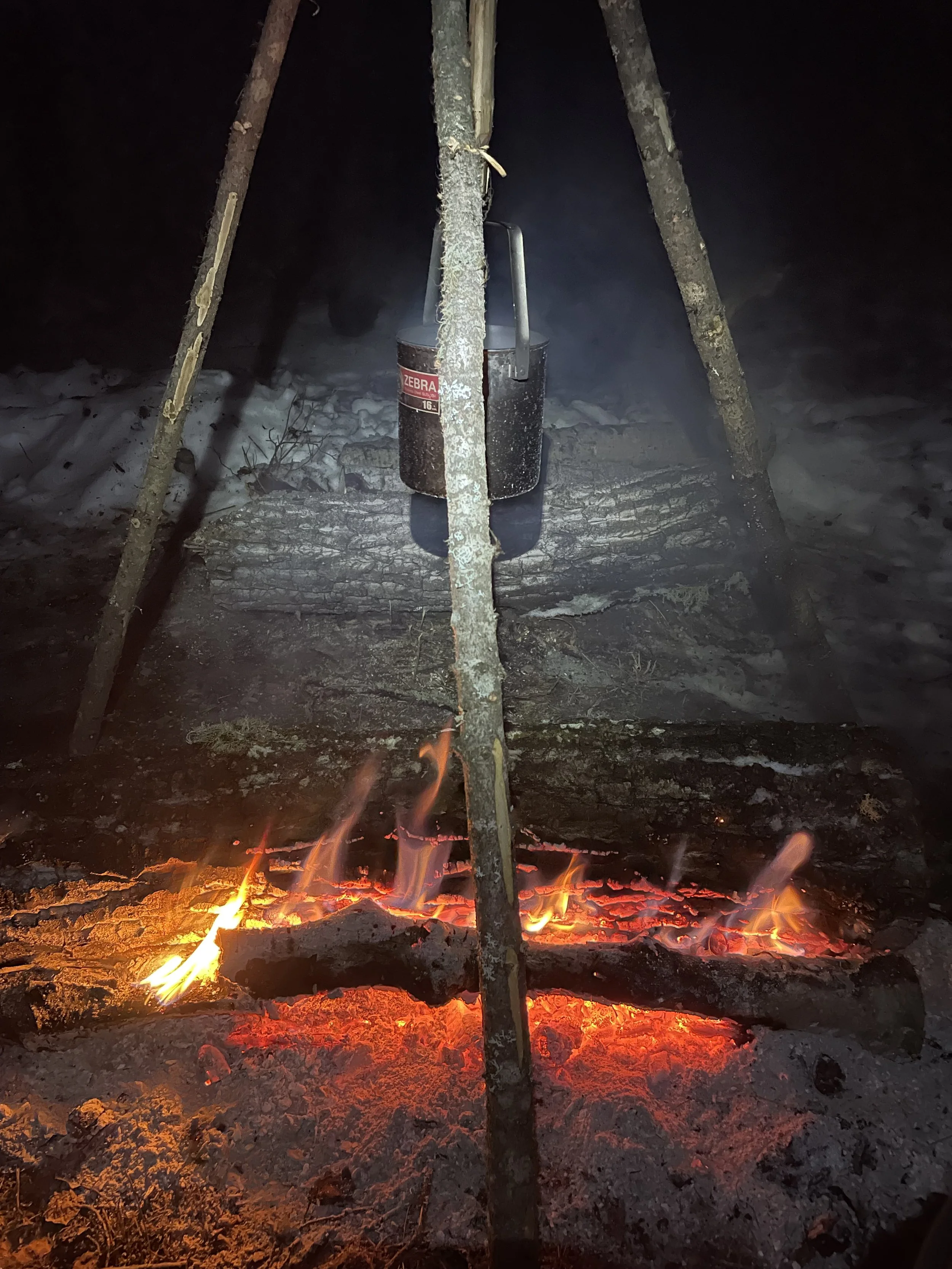 A campfire outdoor at night with a cooking pot suspended over the flames, supported by three sticks.