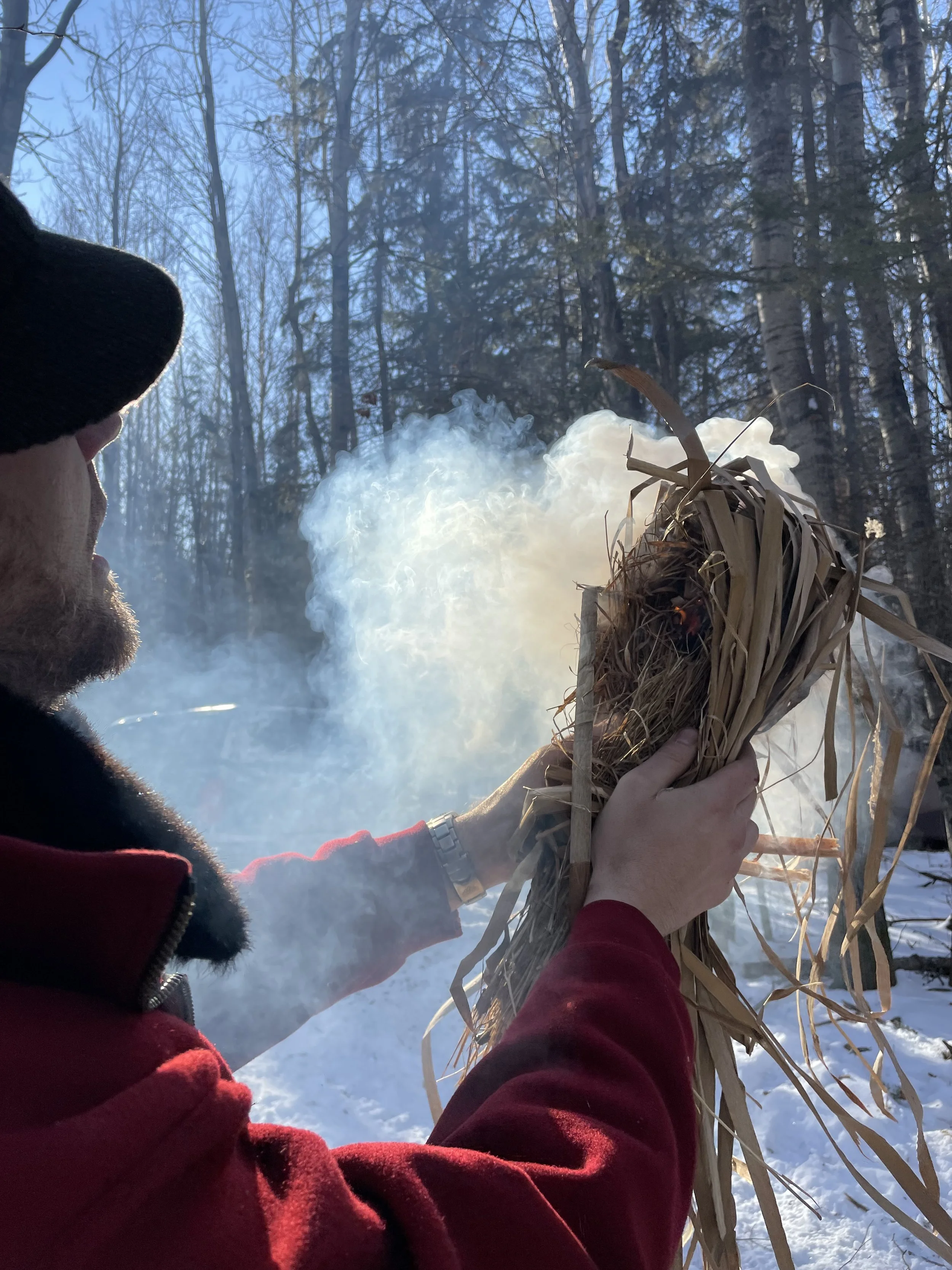 Person holding a bundle of dry grass or reeds over a small fire outdoors in a snowy forest setting.