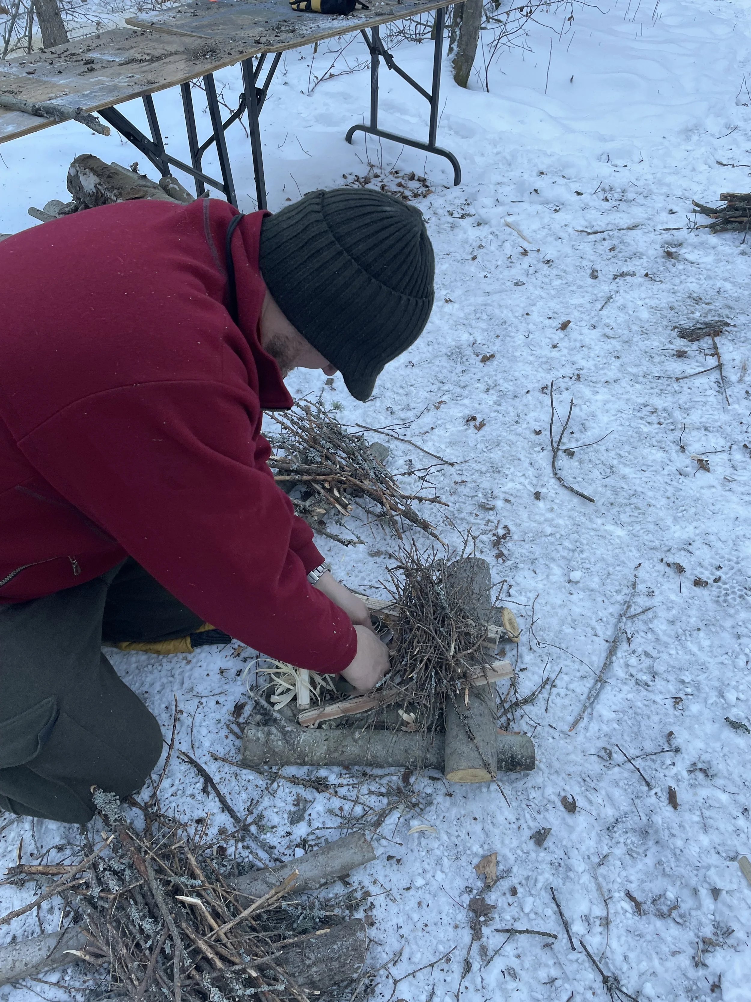 A person wearing a red jacket and black beanie working on assembling a small campfire with twigs and logs on snow-covered ground outdoors.