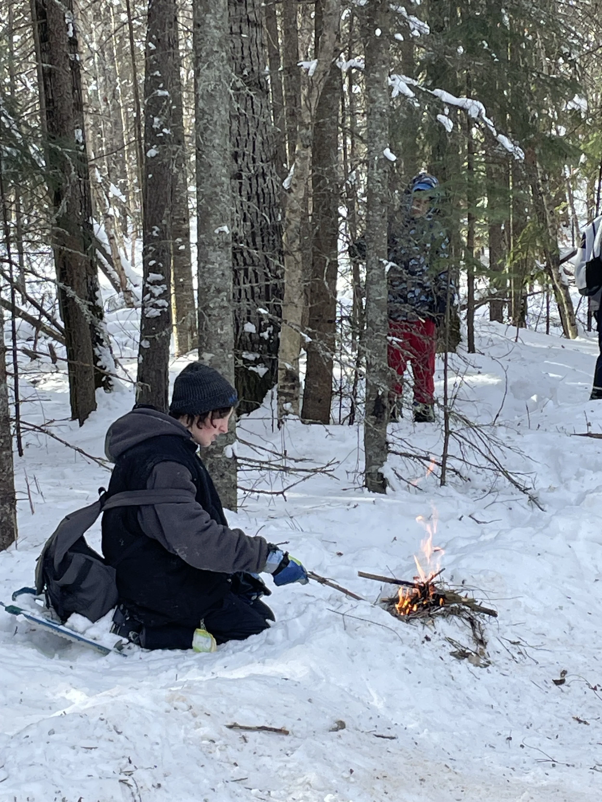 A young person kneeling in the snow, tending to a small campfire, with a backpack and winter clothing, in a snowy forest with several snow-covered trees and two other people observing in the background.