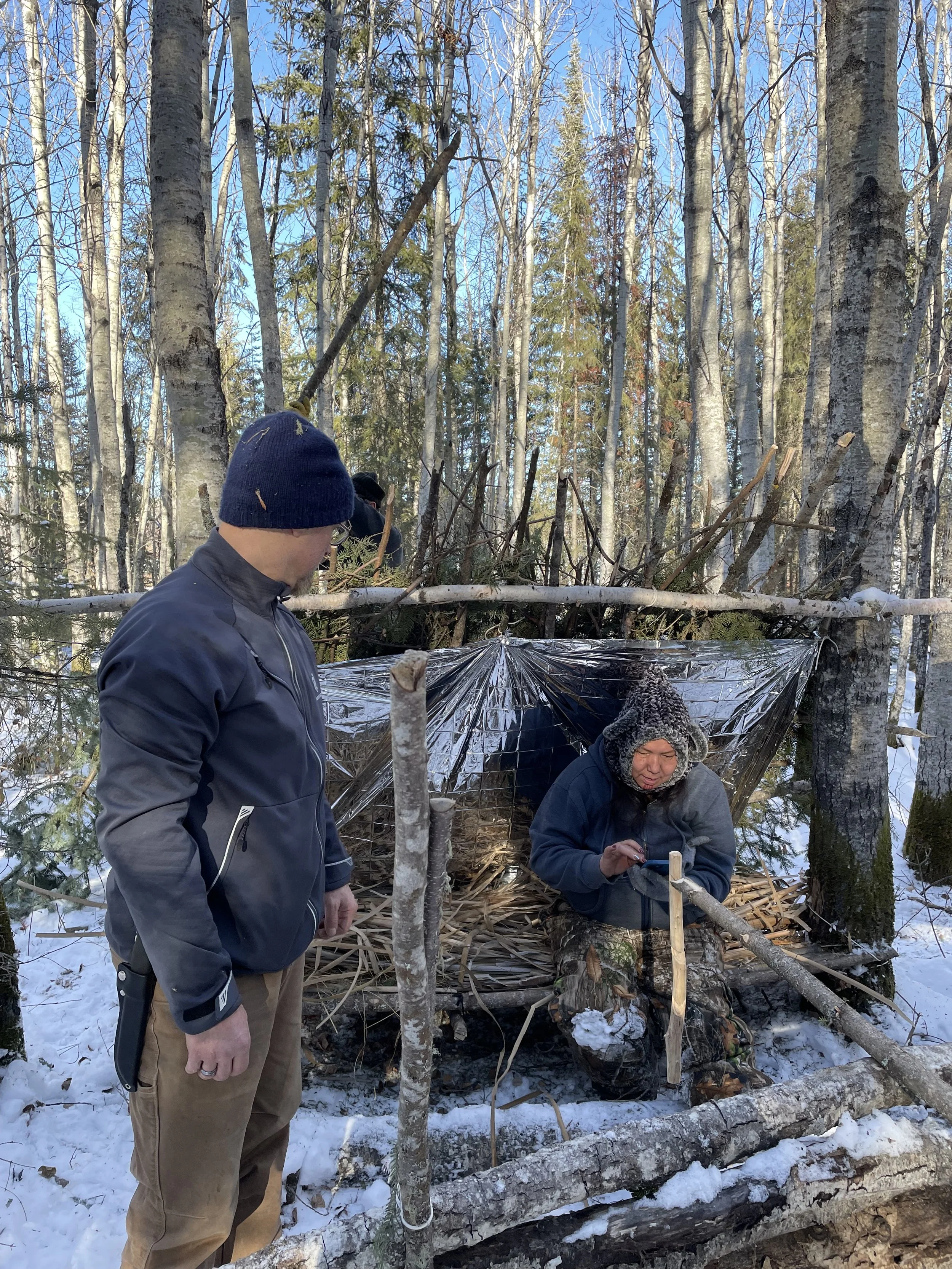 Two men in winter clothing outdoors in a snowy forest, one sitting inside a makeshift shelter made of branches and plastic, the other standing nearby looking at his phone.