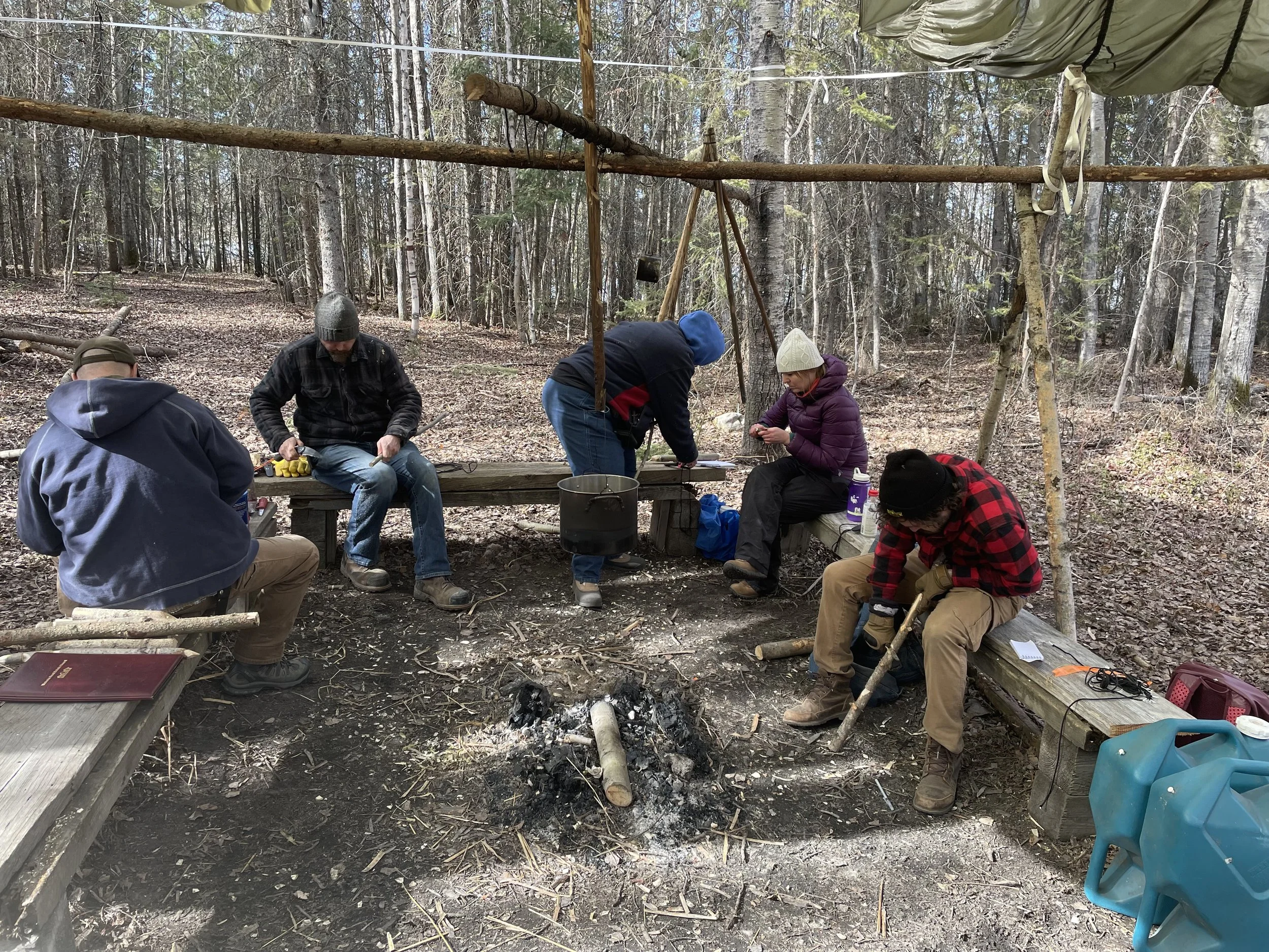 Group of five people sitting and working around a campfire under a tarp in a wooded area, with some tools and supplies nearby.