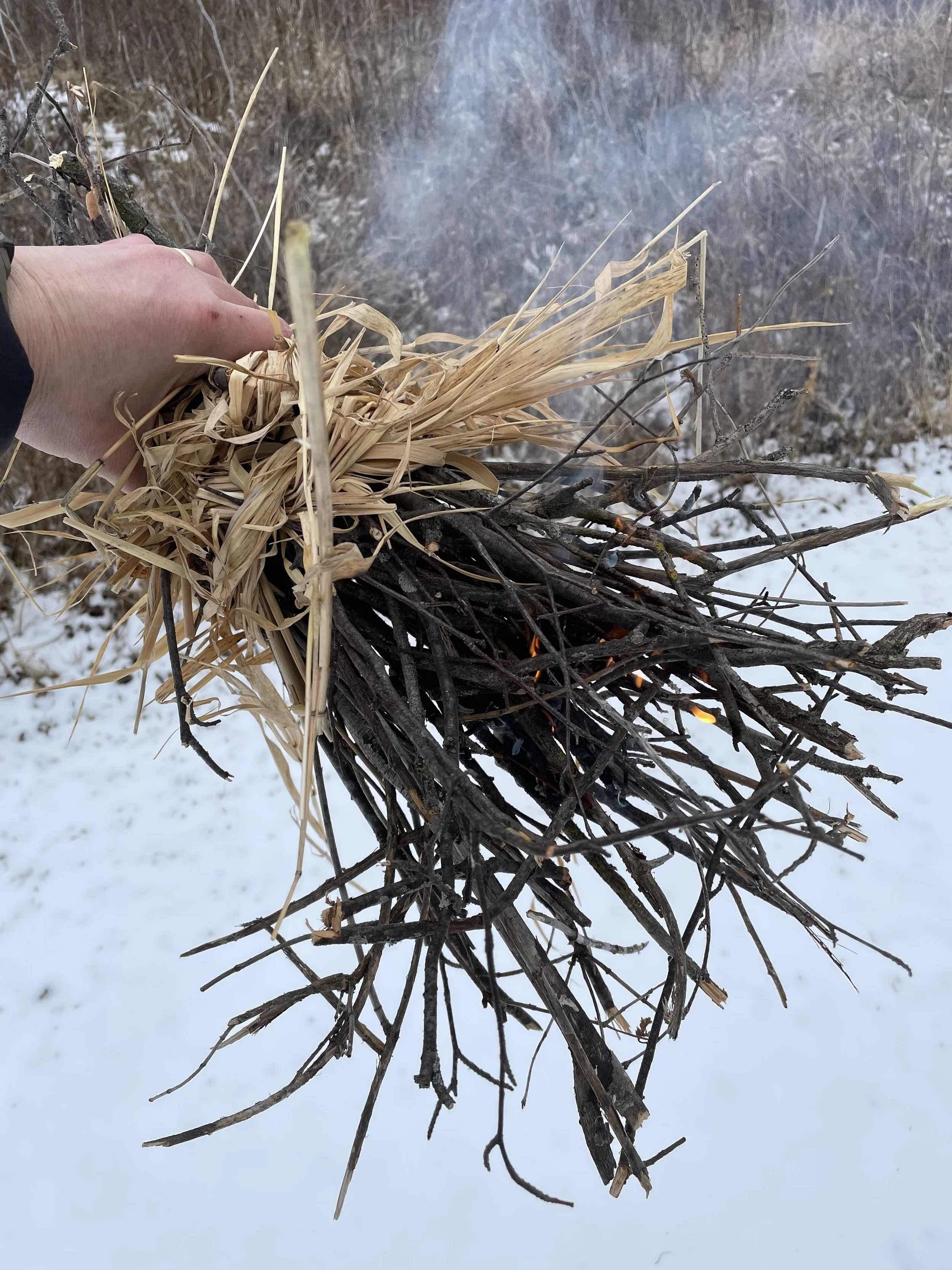 Person holding a bundle of dry sticks and grass over a snowy ground.
