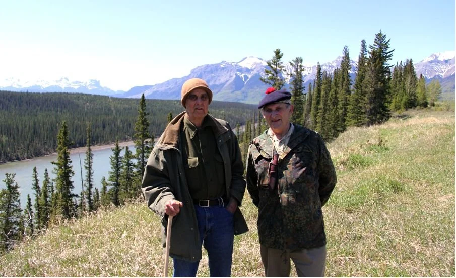 Tom Roycraft and Mors Kochanski in a mountainous, forested area with snow-capped mountains in the background.