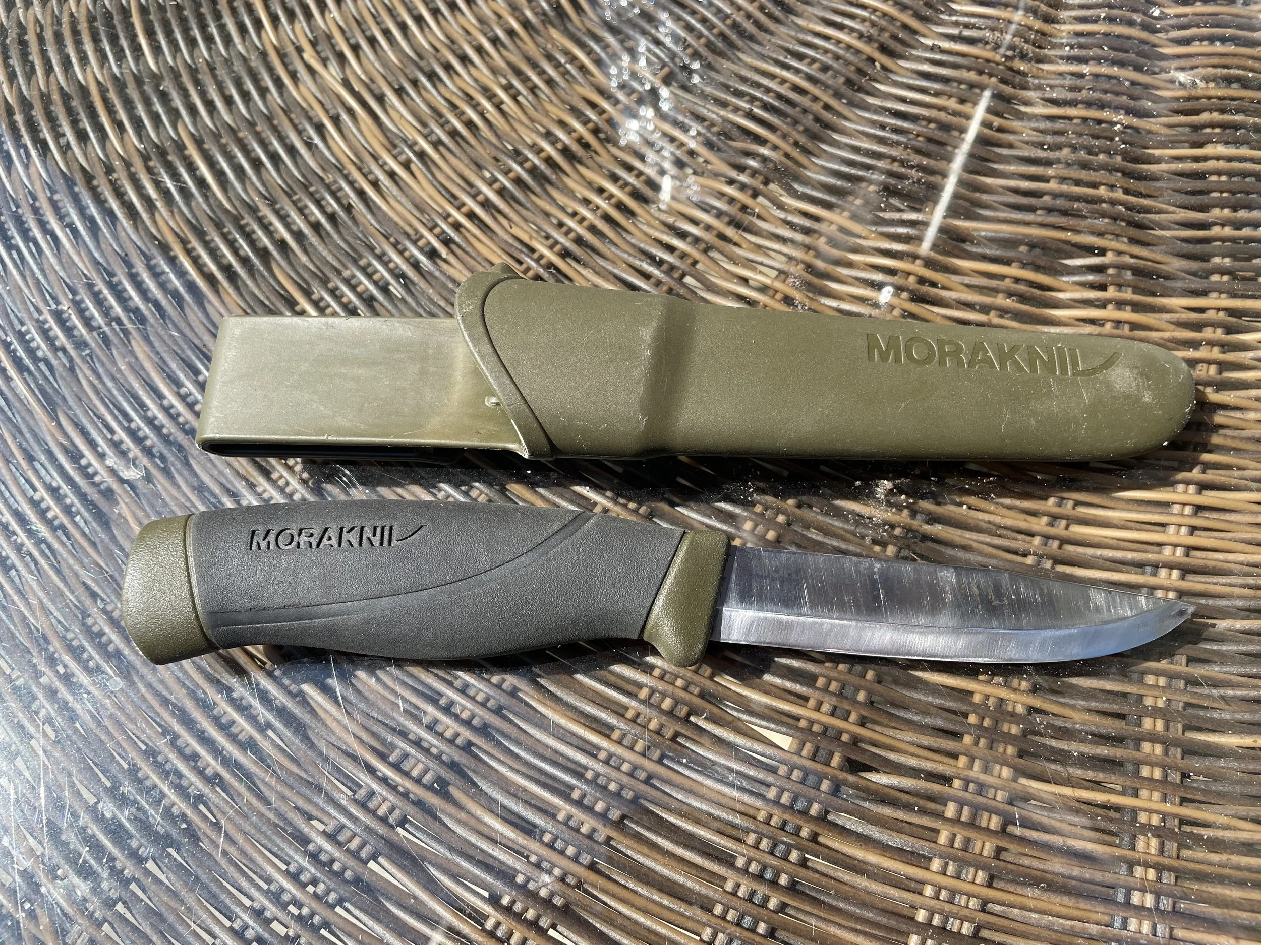 Outdoor hunting or survival knife with a black handle, alongside a green sheath, placed on a woven wicker surface.