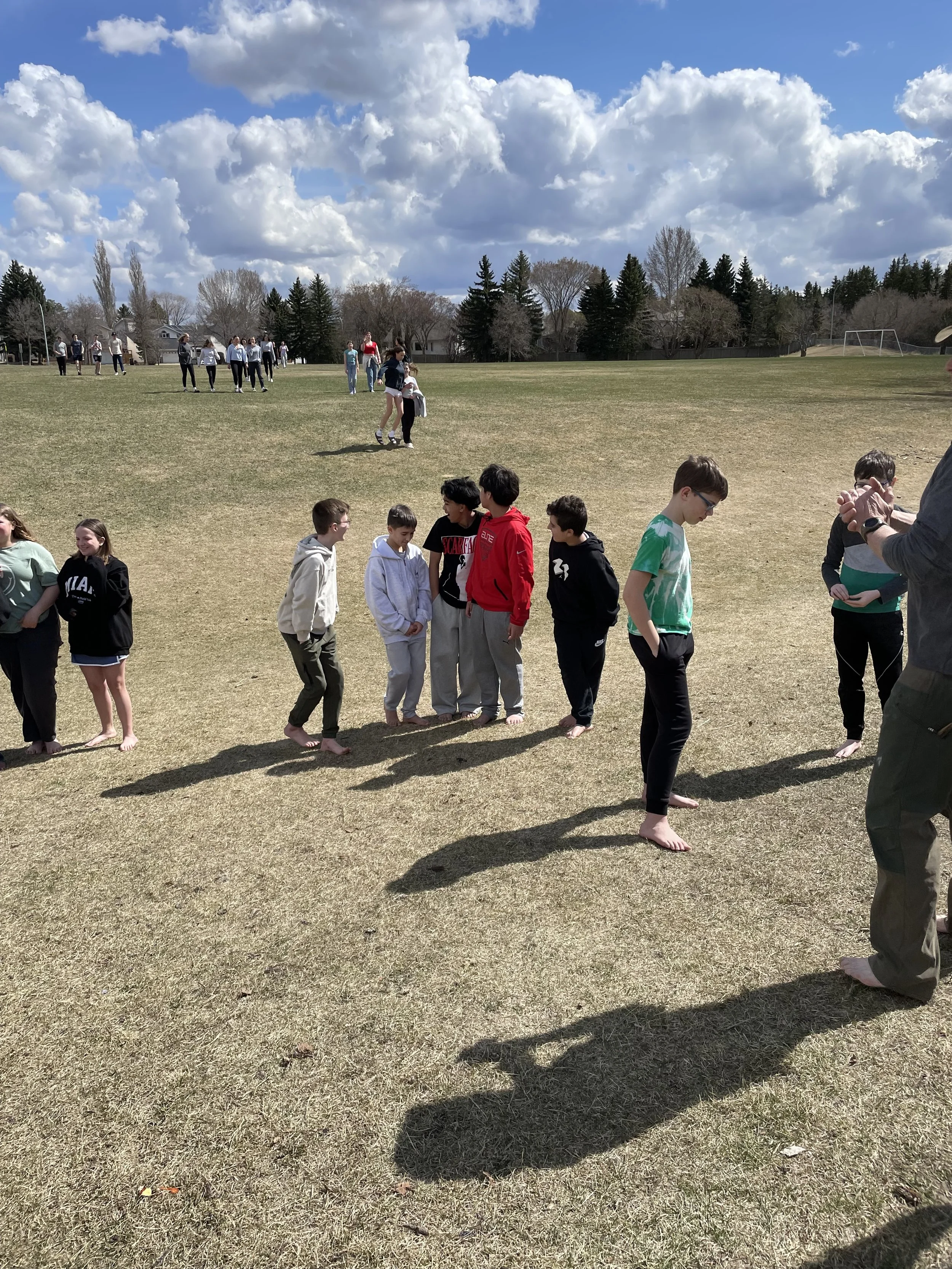Children and adults standing in lines on a grassy field under a partly cloudy sky, participating in an outdoor activity or game.