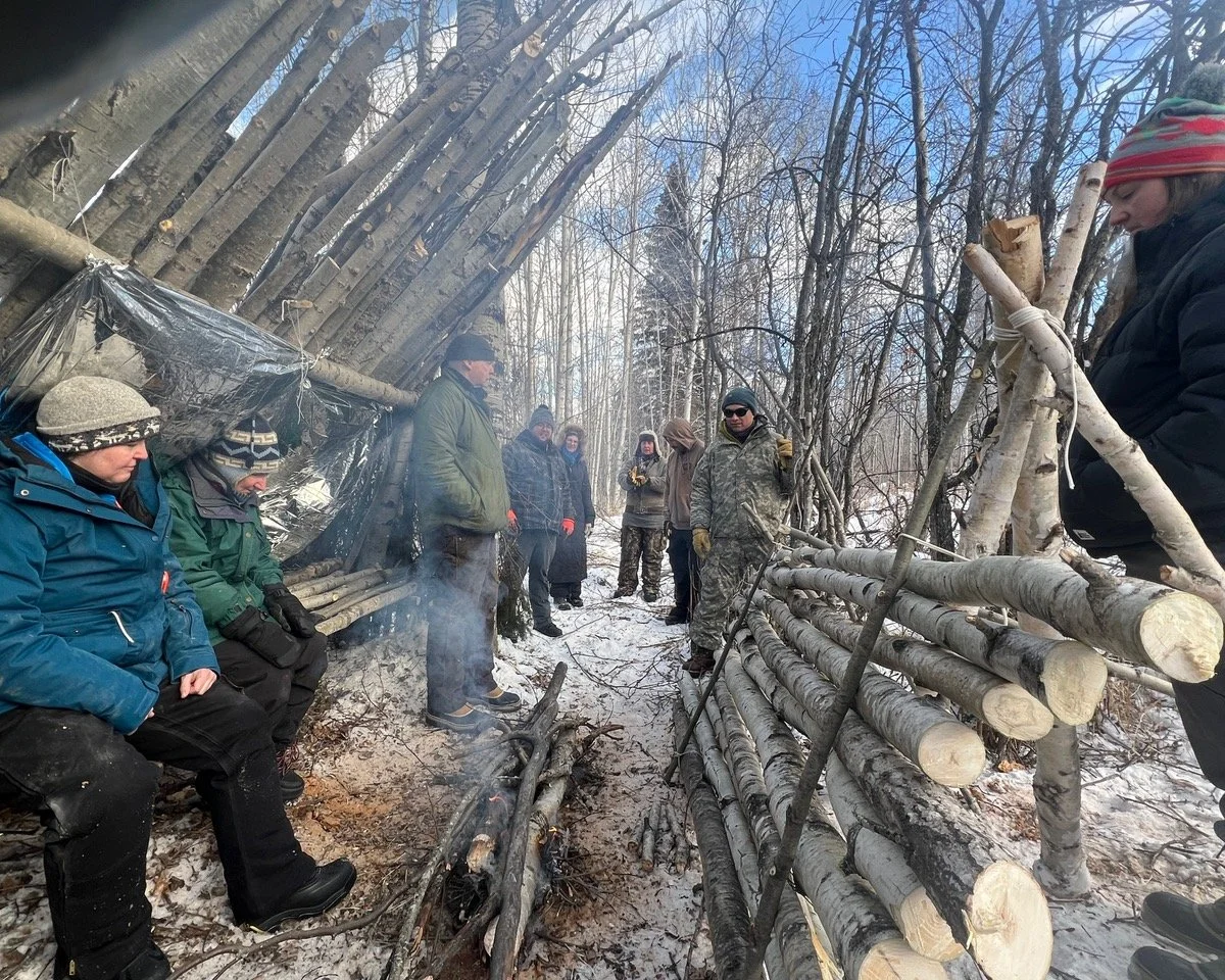 Group of people outdoors in a snowy forest, wearing winter clothing, gathered around a campfire, with a makeshift shelter made of logs and plastic in the background.
