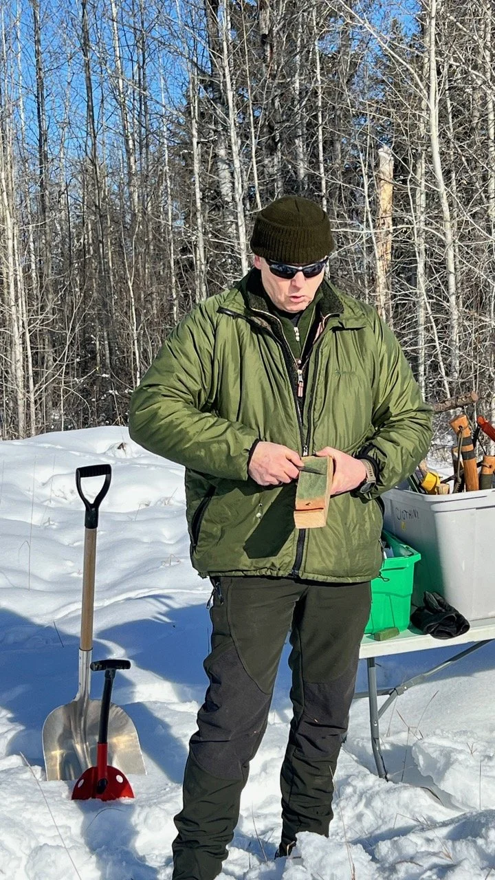 A Instructor in a green winter jacket, black beanie, and sunglasses stands in the snow and demonstrates knife sharpening. There is a shovel and a table with various supplies behind him, with a forest in the background.
