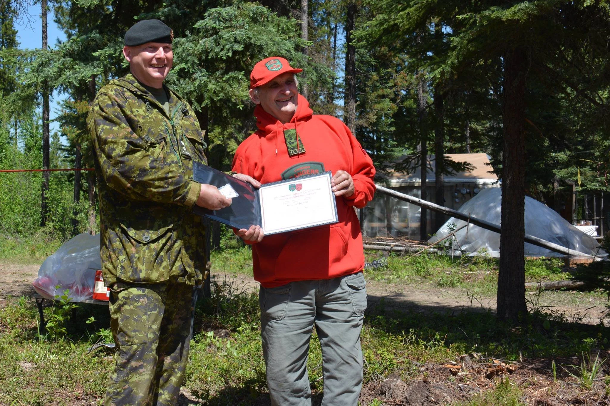 Mors Kochanski outdoors in a forested area, smiling having received a certificate, with trees and some covered structures in the background.