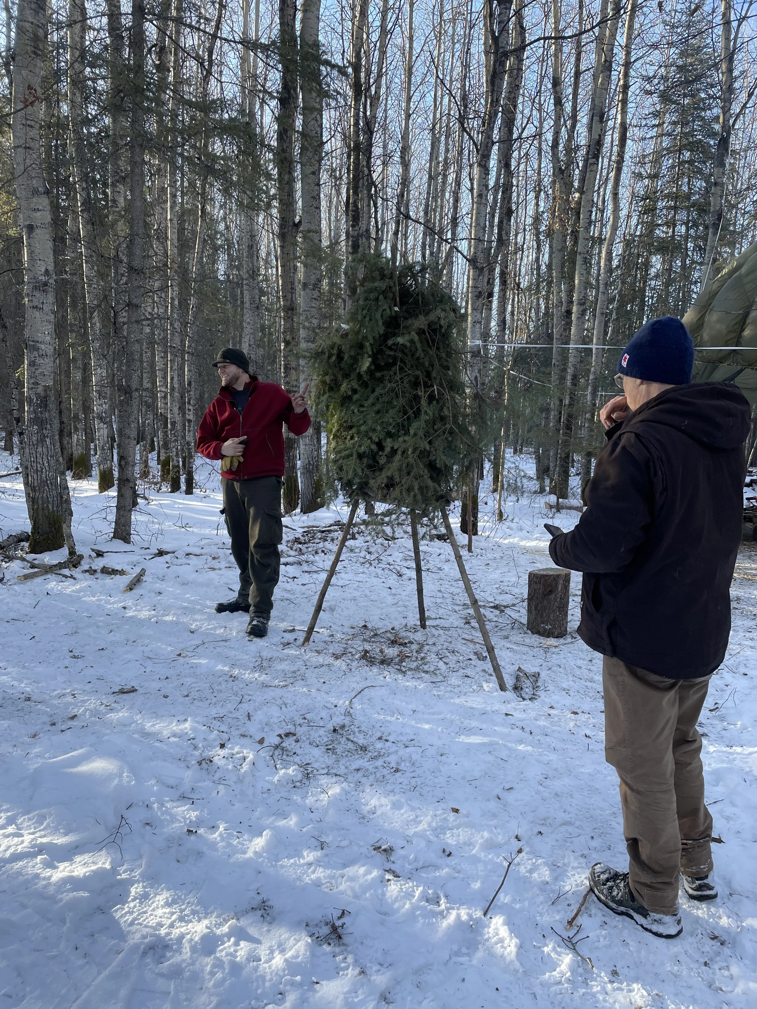 Two men in winter clothing standing in a snowy forest, preparing or inspecting a large Christmas tree set up on a wooden stand.