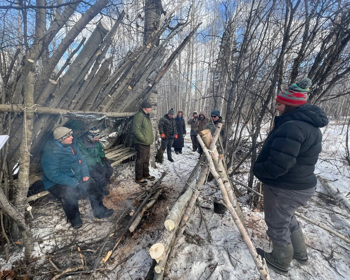 Group of people in winter clothing gathered outdoors in a snowy forest, observing a small shelter made of fallen branches and a tarp against trees.