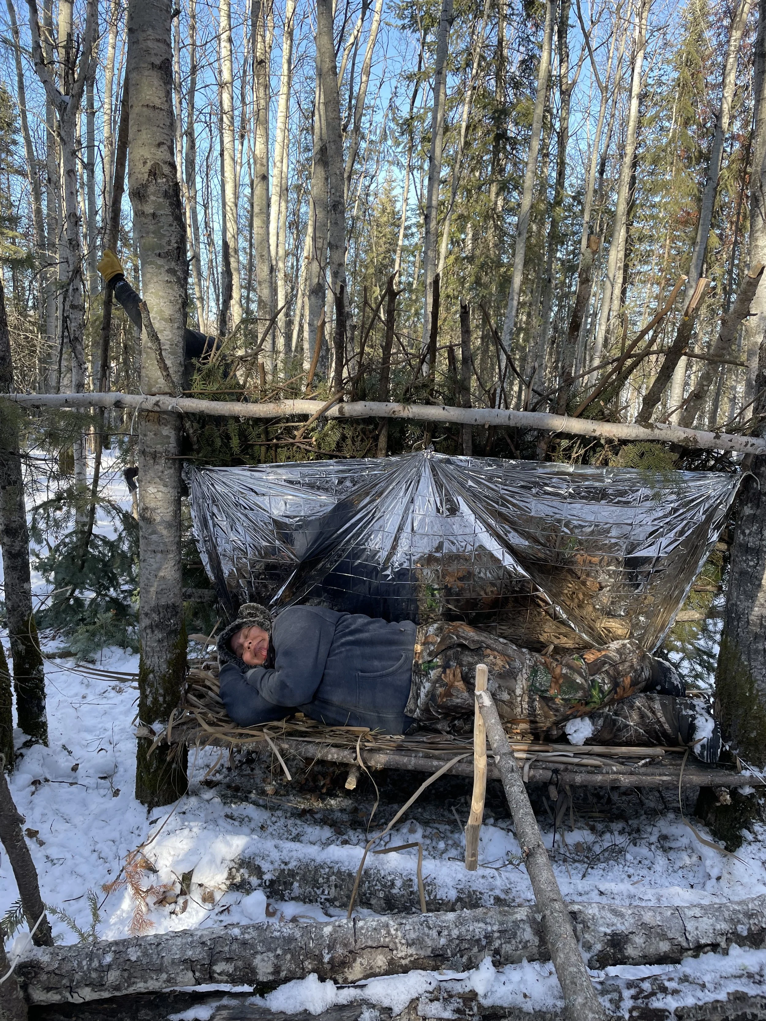 A man lies on a makeshift bed in a forest, covered with a camouflage blanket, with a reflective emergency blanket behind him, during winter. He is smiling and resting in the snow.