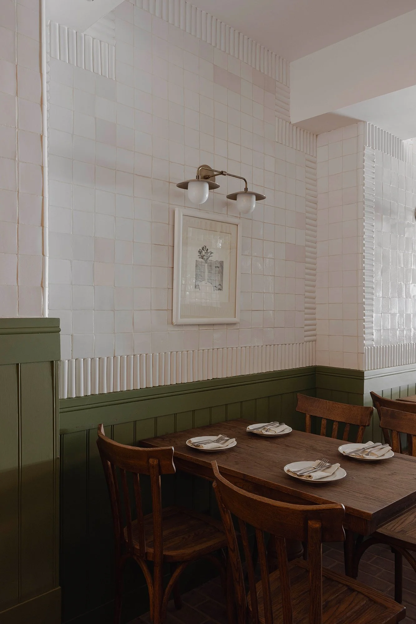 Wood dining table with vintage chairs set for four, against a white handcrafted tile wall with vertical trim detail and olive green wainscoting, accented by a brass sconce and framed botanical artwork at The Lamb restaurant in Mexico City