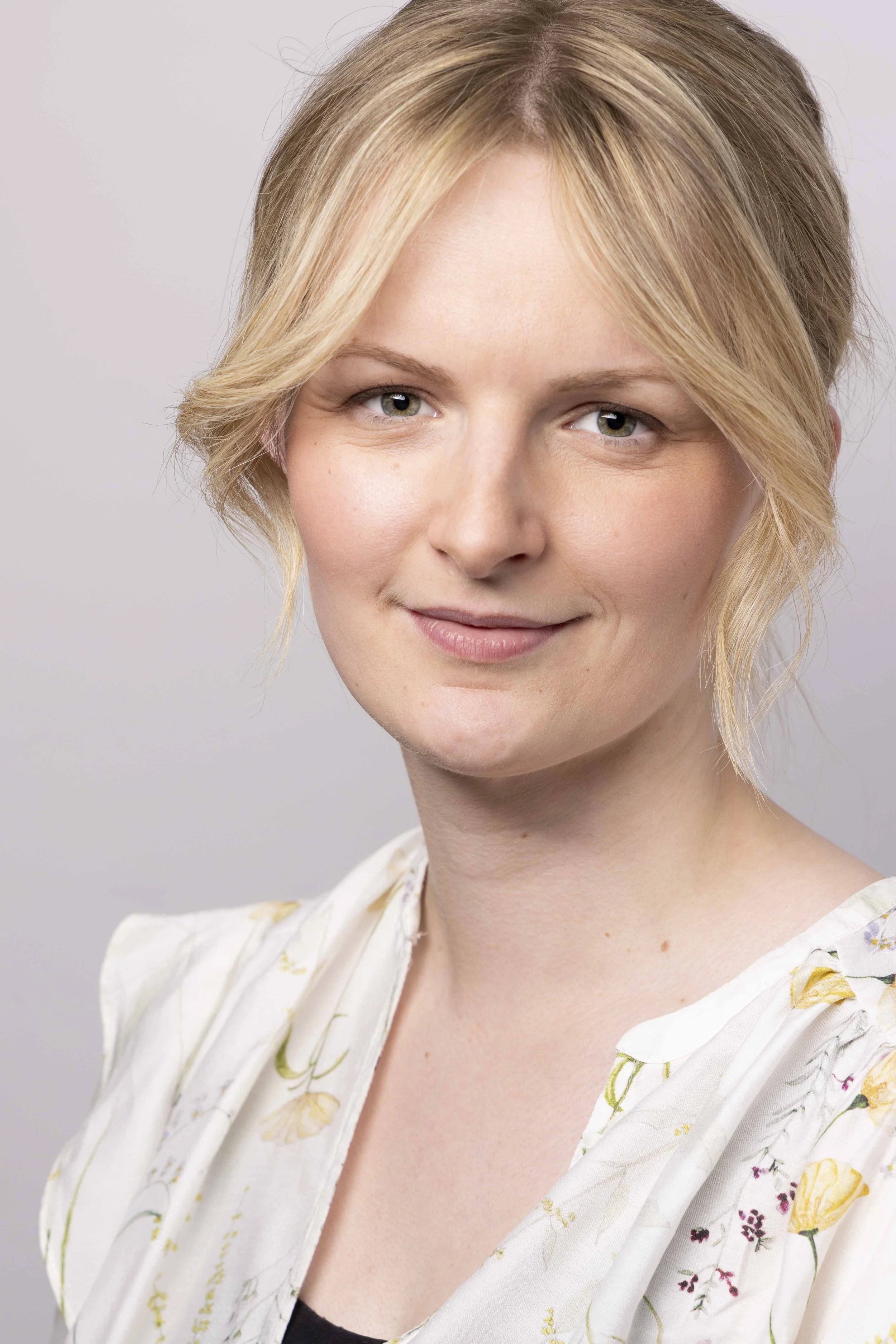 A woman with short blonde hair smiling softly, wearing a light-colored floral blouse, against a plain light background.