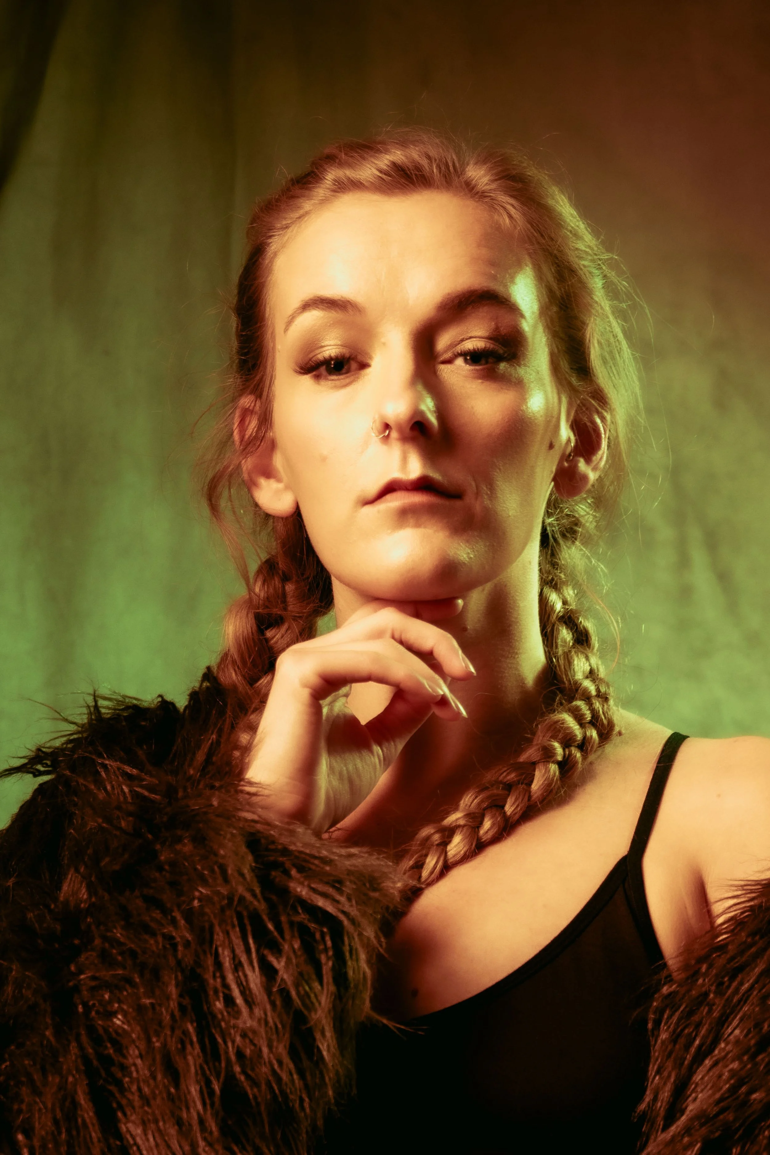 A young woman with braided hair, wearing a black spaghetti strap top and a fur coat, poses with her hand resting on her chin, against a green backdrop with yellow lighting.
