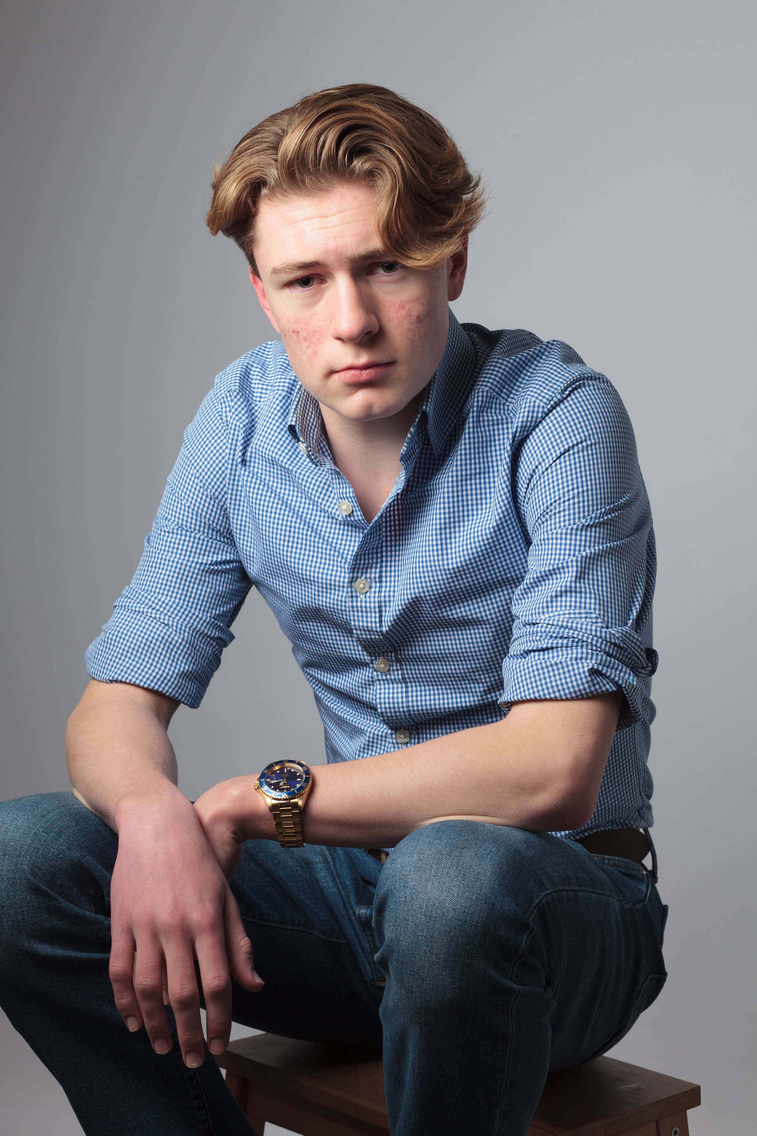 A young man with light skin, reddish-brown hair, and facial acne, wearing a blue checkered shirt with rolled-up sleeves, dark jeans, and a gold watch, sitting on a wooden stool against a light gray background, looking at the camera with a serious expression.