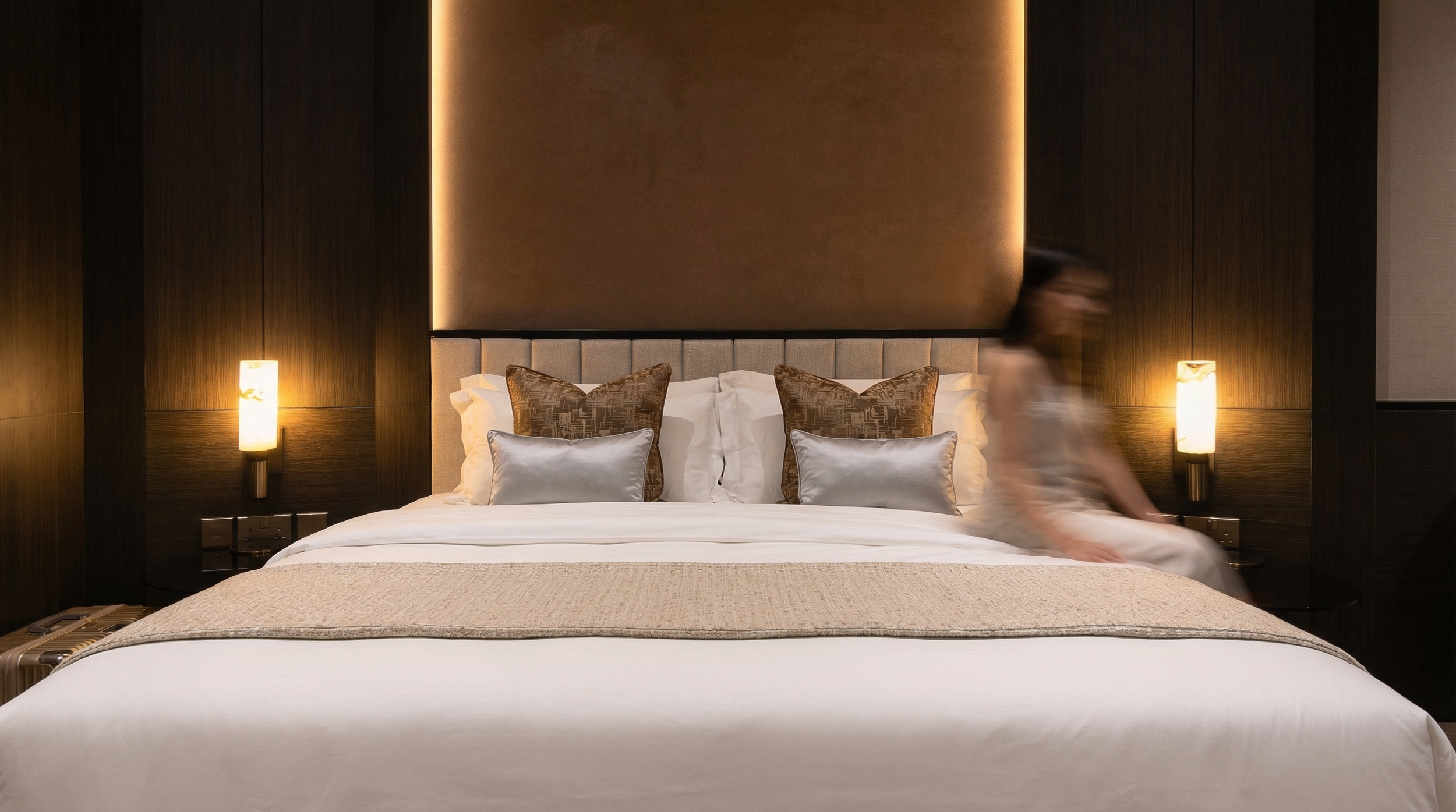 A neatly made bed with white linens, decorative pillows, and a beige bed runner, set against a dark wood-paneled wall with two wall-mounted lamps on either side, and a woman sitting on the right side of the bed.