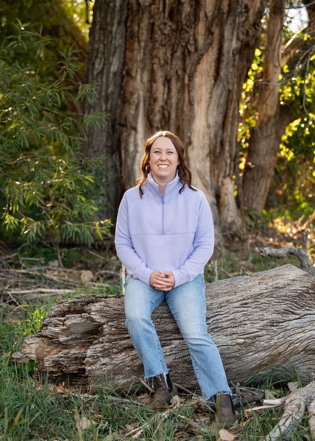 Sarah Henry, National Board-Certified Health and Wellness Coach, sitting outdoors on a fallen tree in a lavender pullover.