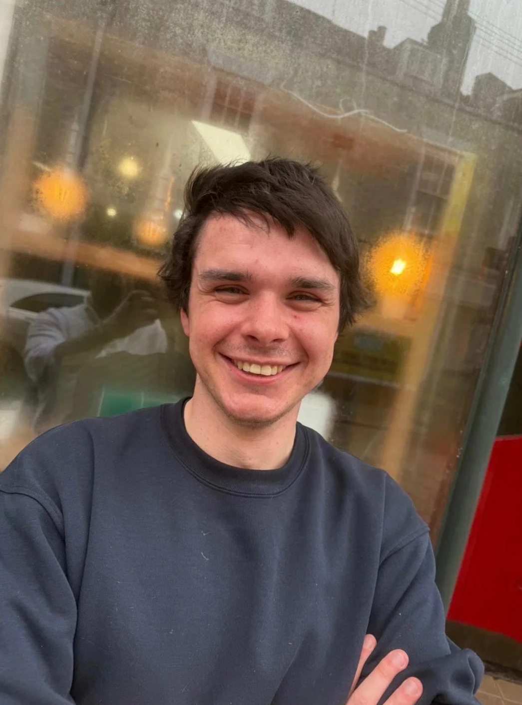 A young man with dark, wavy hair and a big smile taking a selfie outside near a glass window with reflections of city buildings and warm lights in the background.