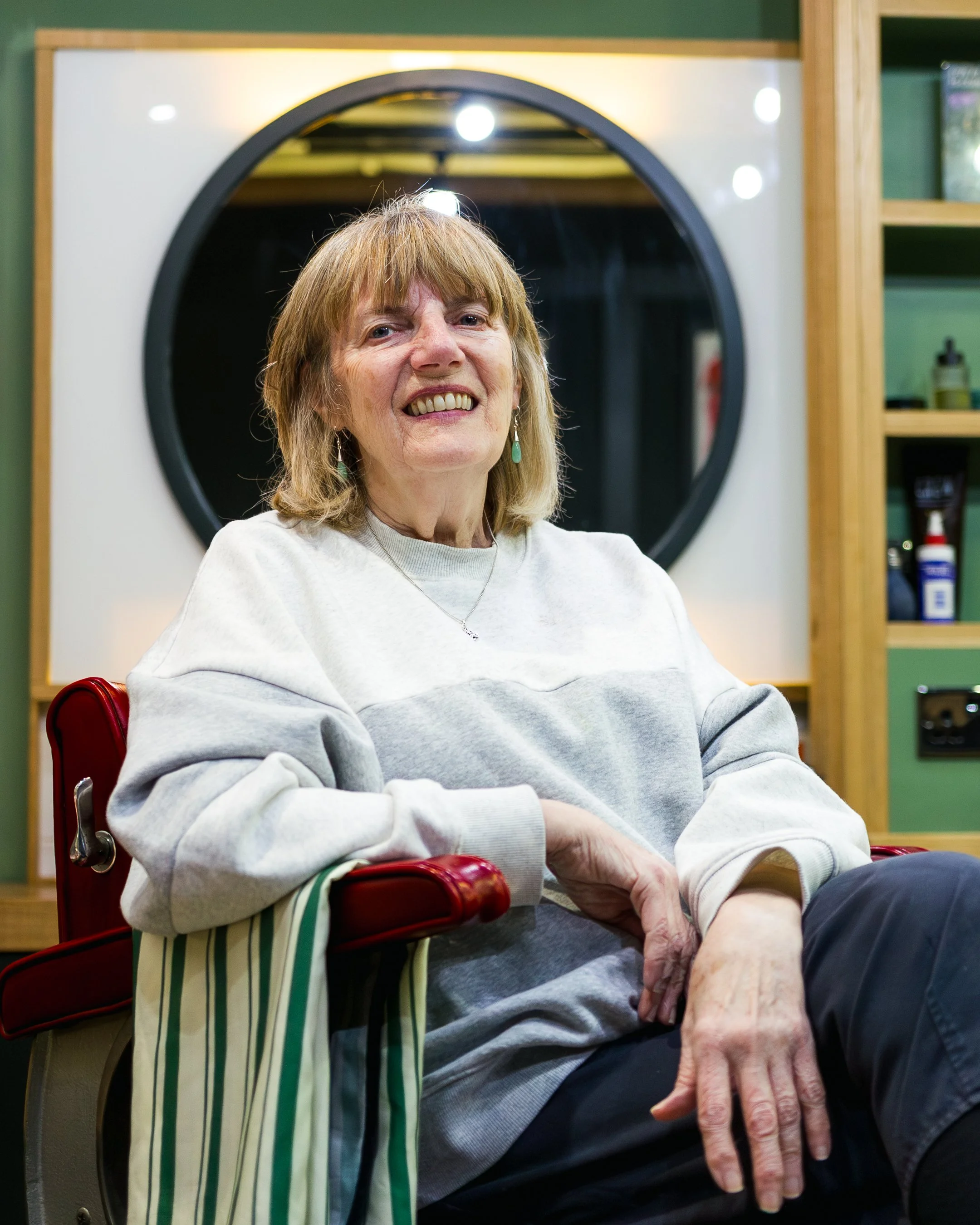 An elderly woman with short blond hair and earrings sitting in a red chair, smiling, in a room with green walls and wooden shelving, wearing a light gray sweatshirt and black pants.