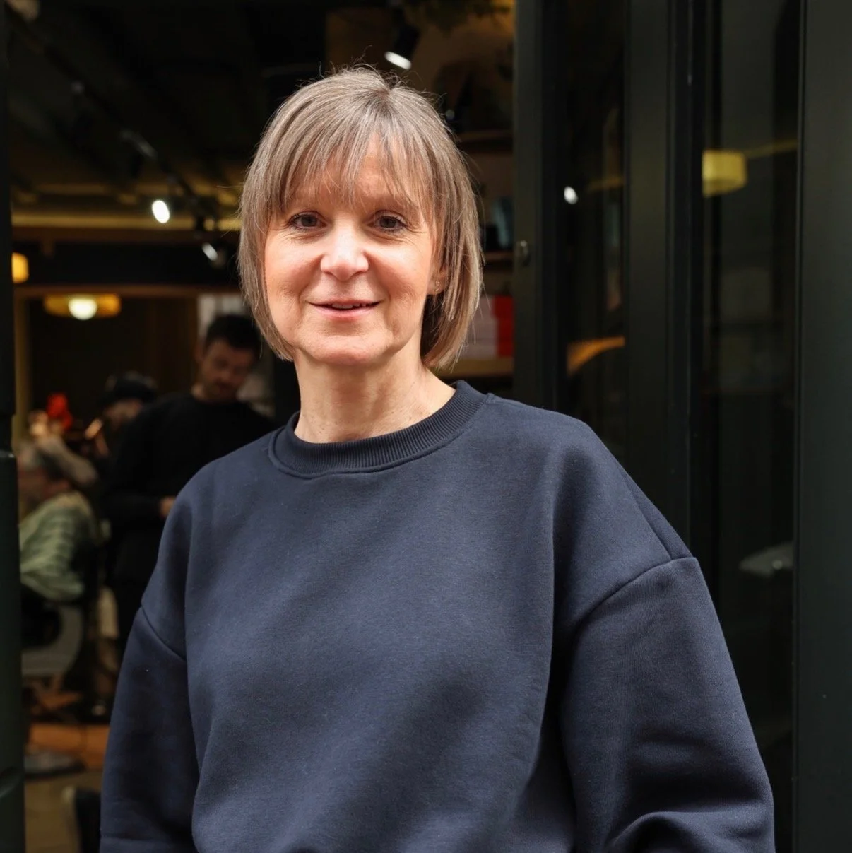 A middle-aged woman with short light brown hair wearing a navy blue sweatshirt, standing indoors with a blurred background of a restaurant or cafe environment.