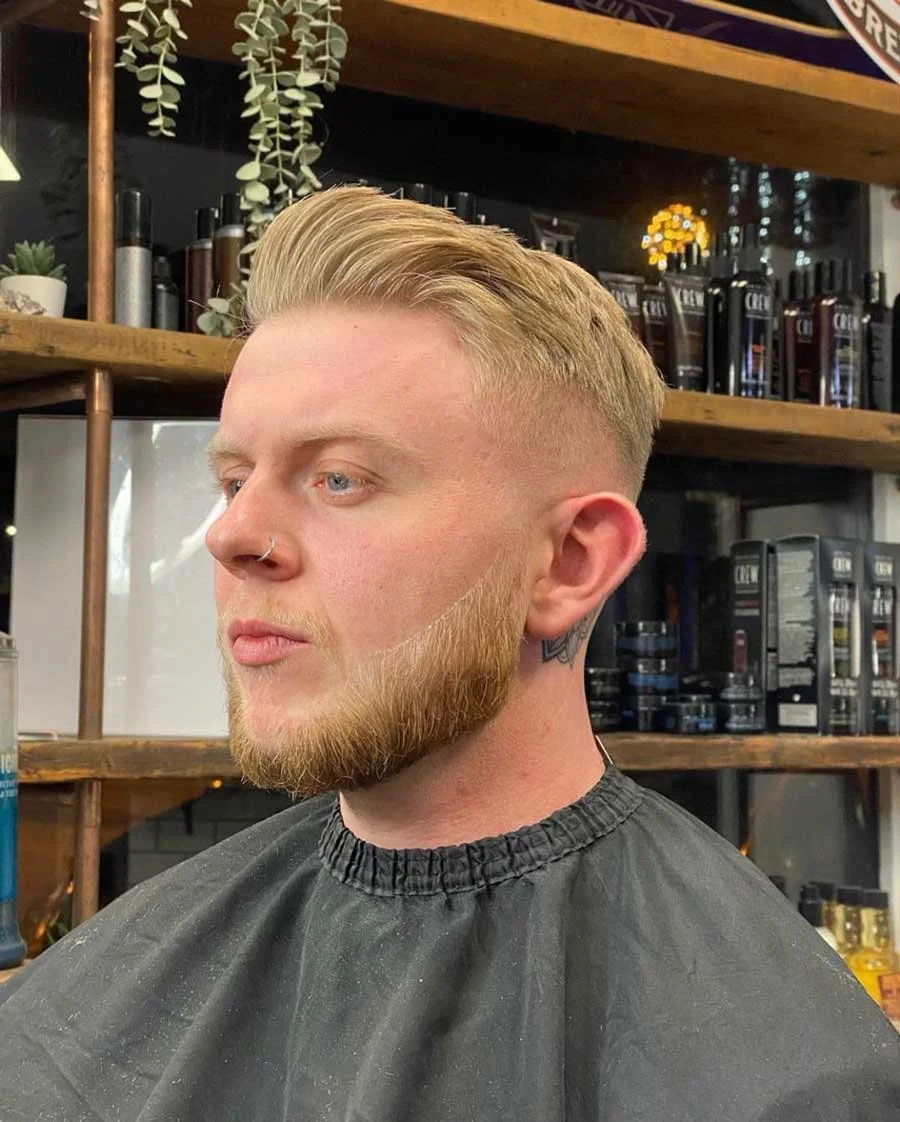 A man with blond hair and a full beard sitting in a barber's chair in a barbershop, with shelves of grooming products in the background.