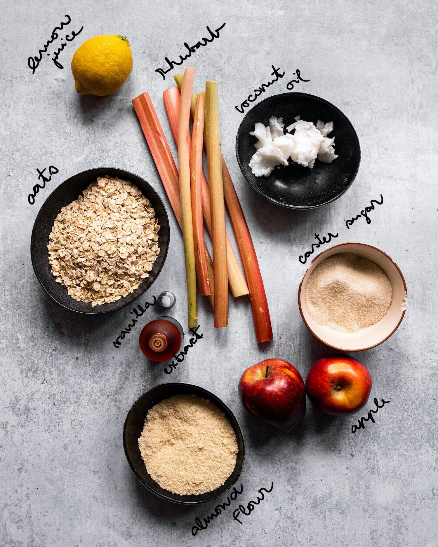 All ingredients that are needed for rhubarb crumble - rhubarb, lemon juice, almond flour, oats, apple, vanilla extract, caster sugar and coconut oil.