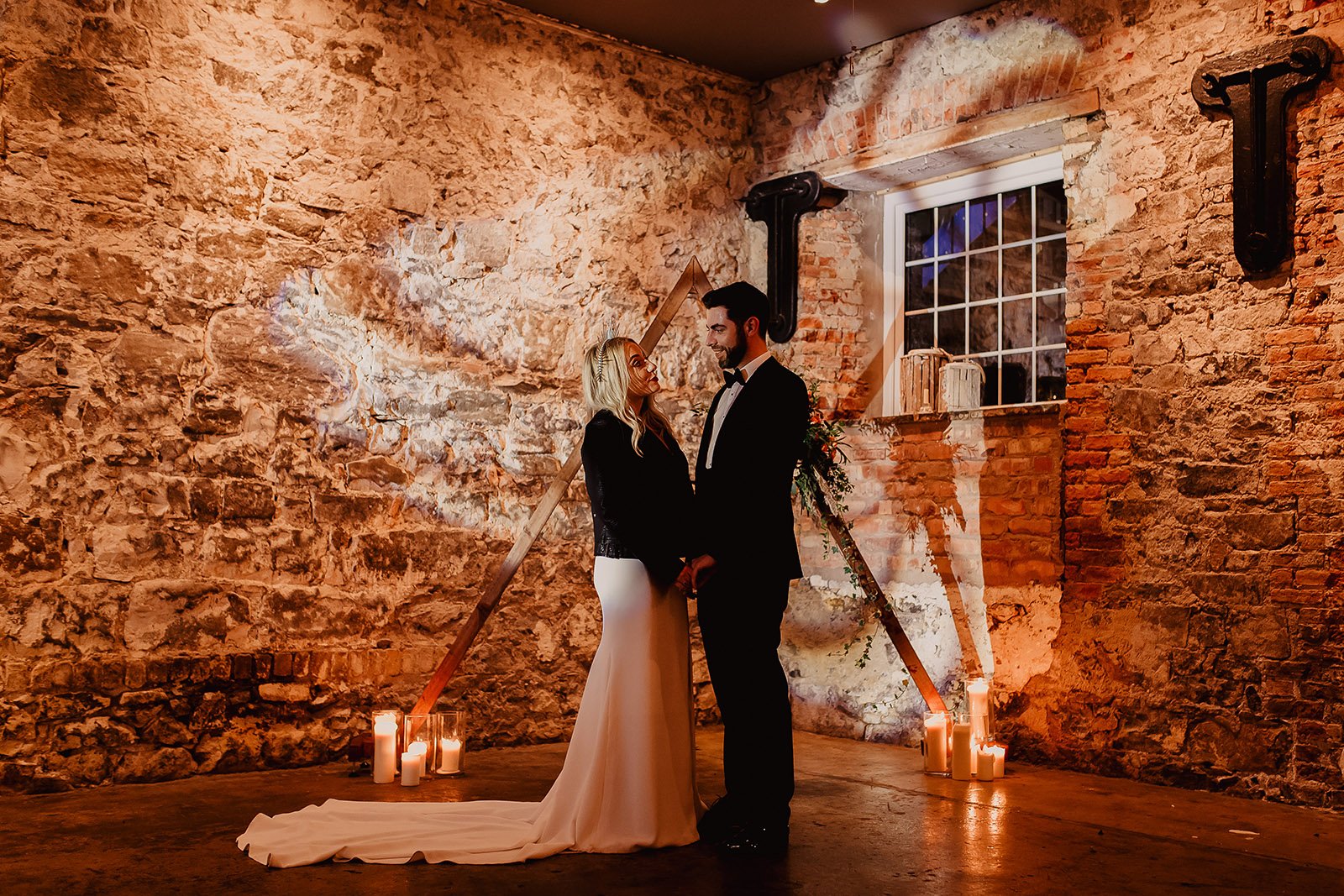 a bride wearing a long white dress and black leather jacket holds hands with and looks at her groom in a green velvet jacket as they stand in front of a candle lit triangular wedding arch with lights from the dj booth casting a heart shape on the red