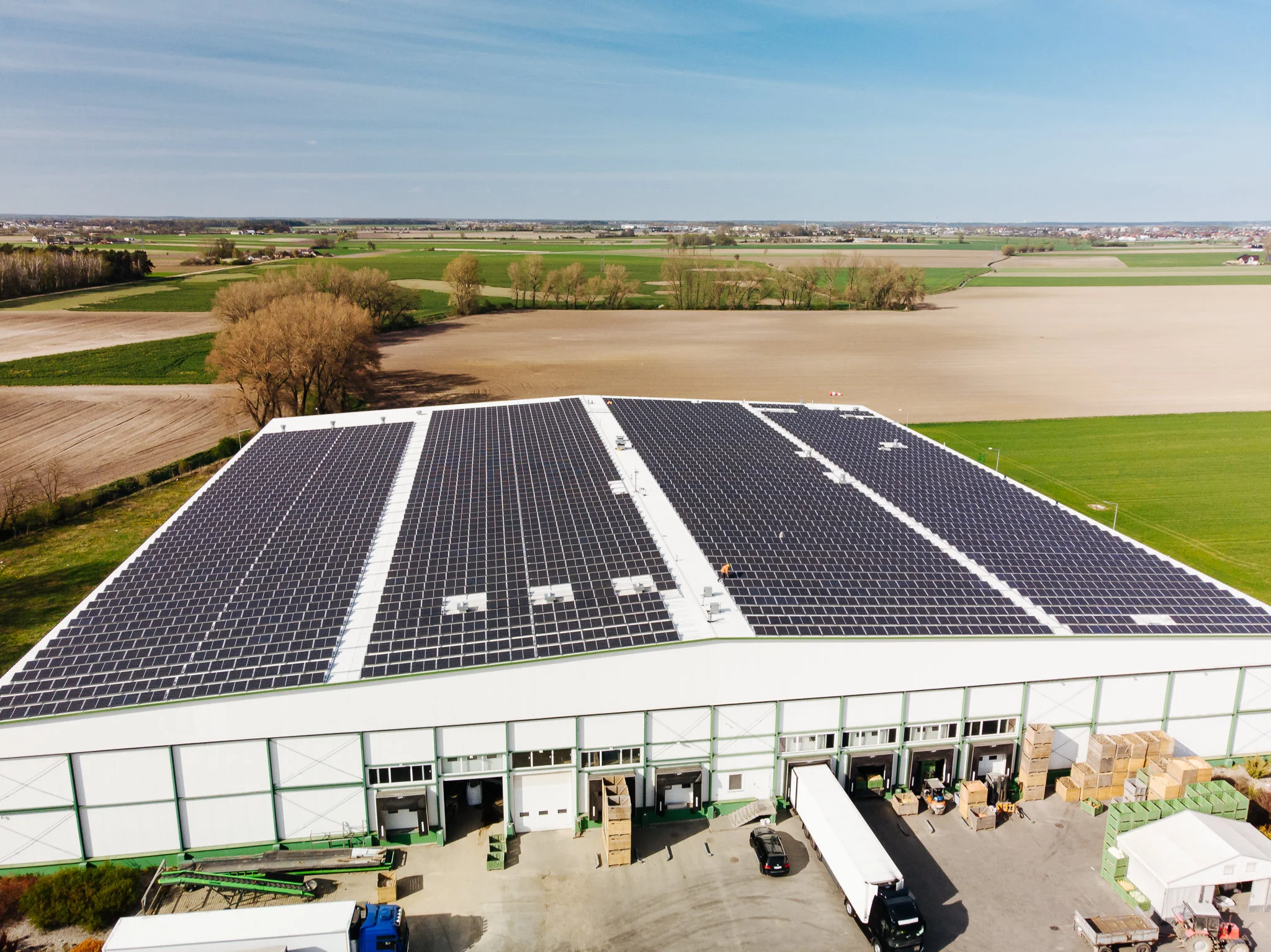 Aerial view of an industrial warehouse with a large solar panel array on the roof, surrounded by fields and farmland.