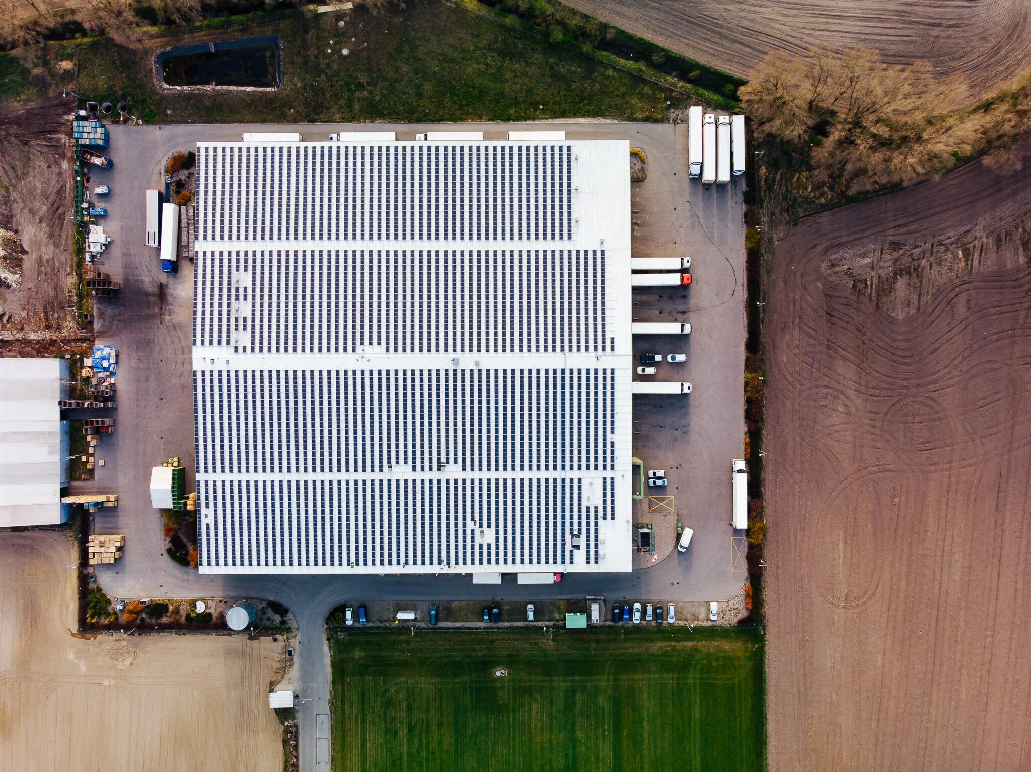 Aerial view of a large building with solar panels on its roof, surrounded by parking lots with trucks and cars, fields, and patches of trees.