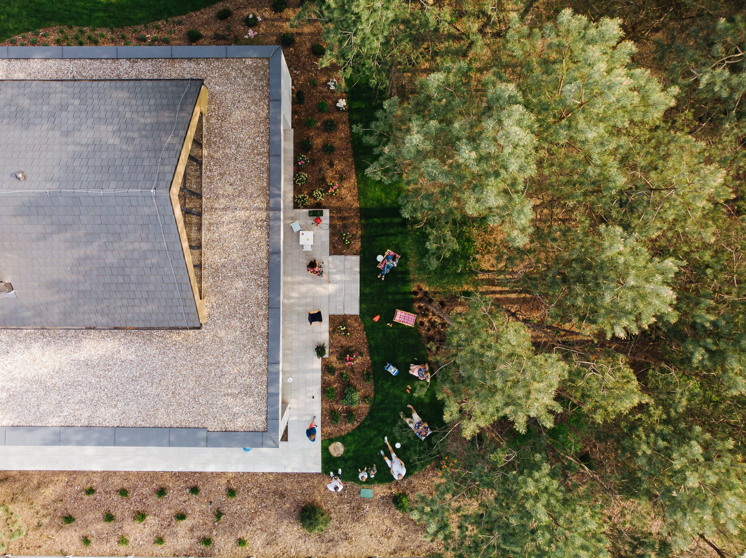 An aerial view of a backyard with a house, green lawn, trees, and people sitting and standing on the grass.