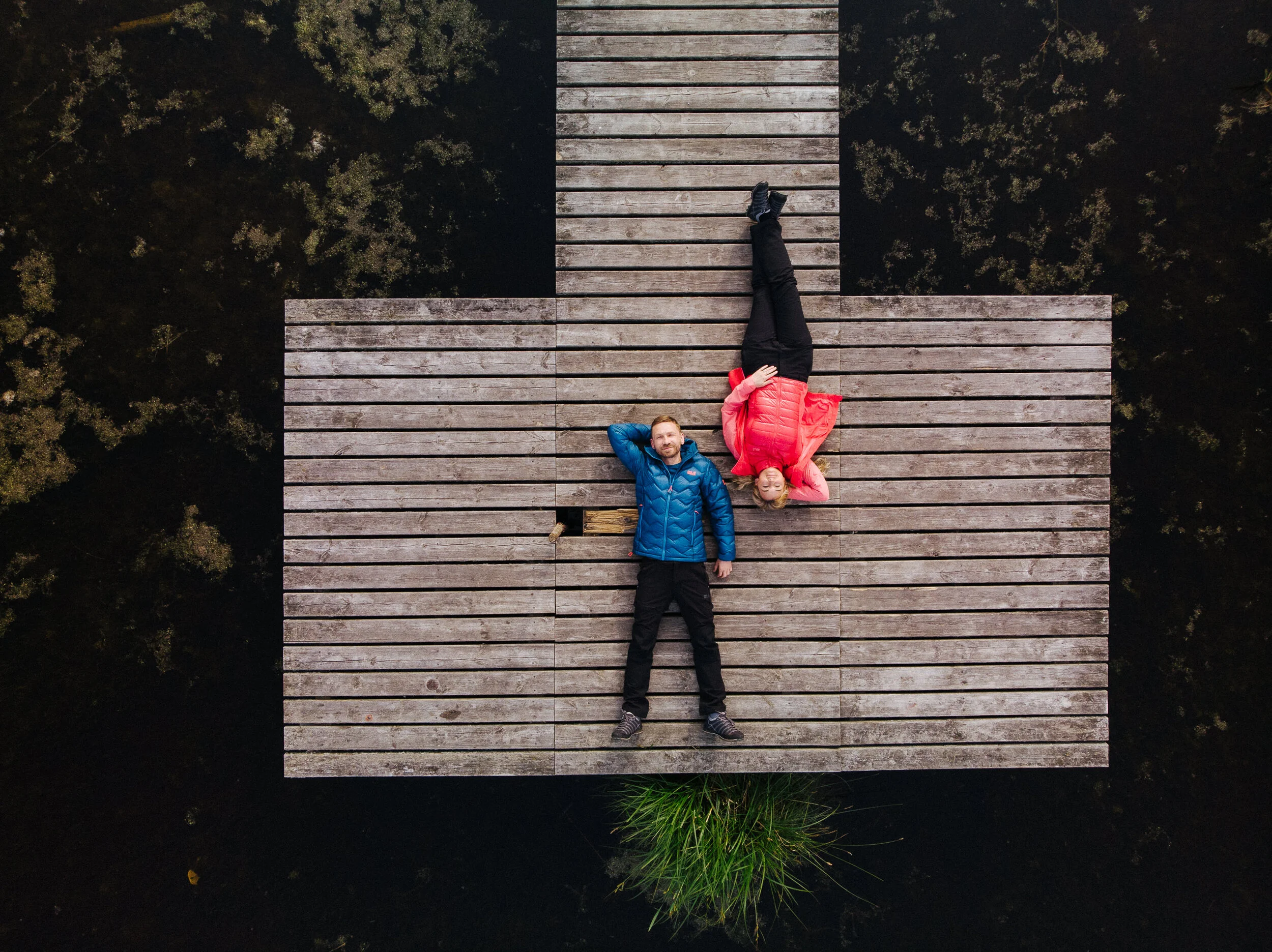 Two people lying on a wooden dock over dark water, seen from above. The man is wearing a blue jacket and black pants, lying with hands behind his head. The woman is wearing a bright pink jacket and black pants, lying on her side with her head resting