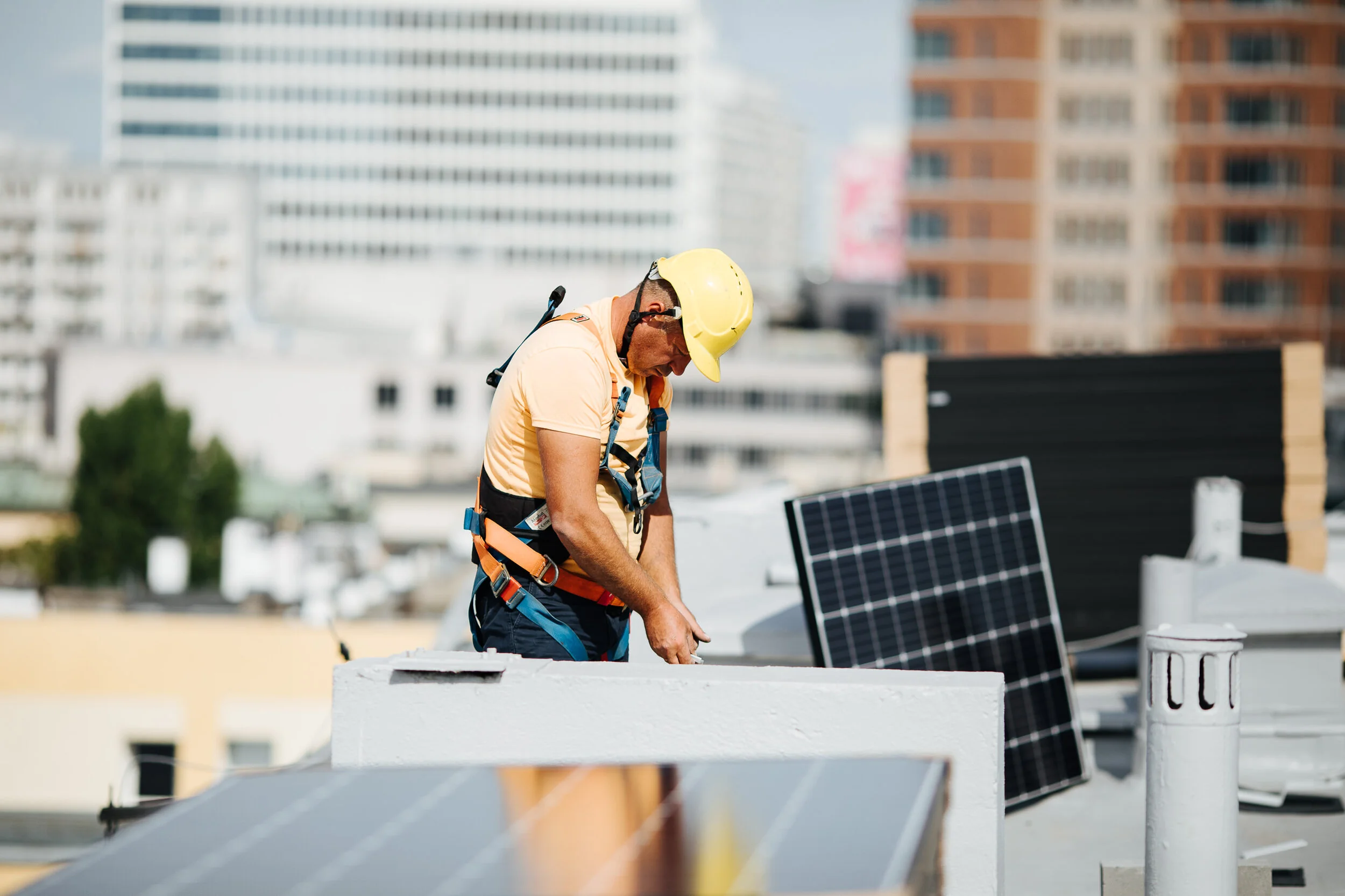 A construction worker wearing a yellow hard hat and safety harness working on the roof of a building, with solar panels nearby and city buildings in the background.