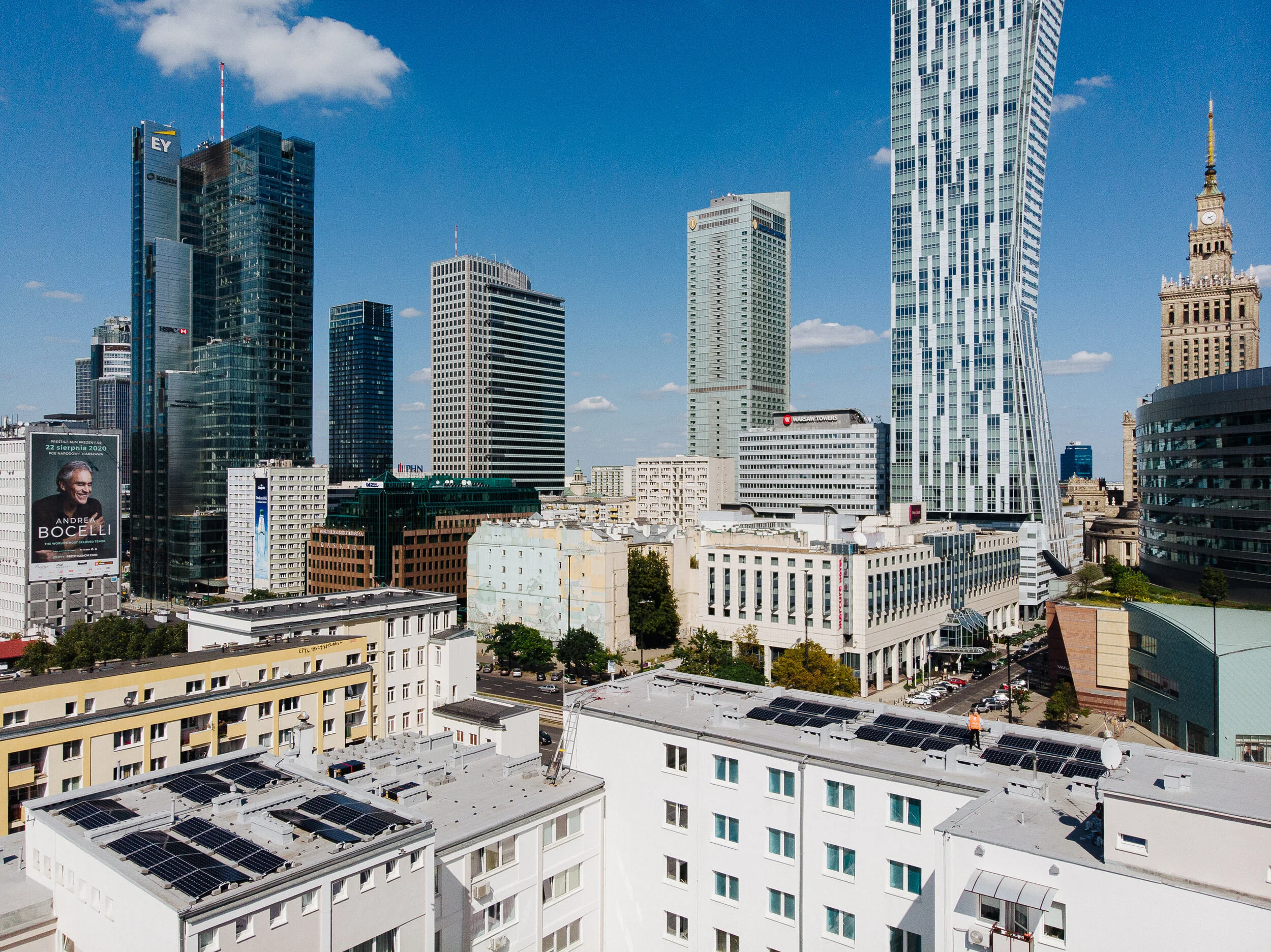 Cityscape of high-rise buildings and skyscrapers in downtown, with clear blue sky and scattered clouds, featuring modern architecture and solar panels on some rooftops.