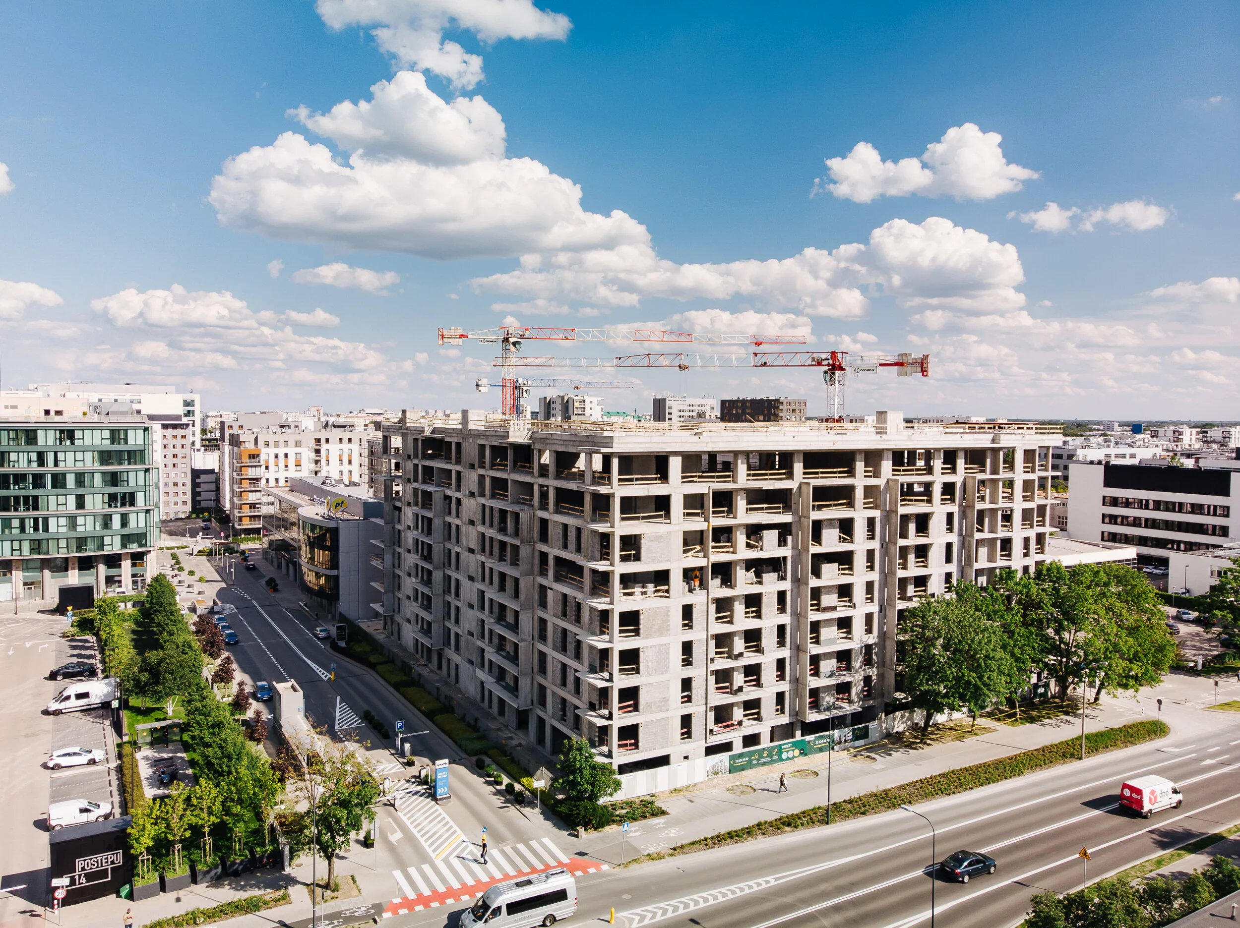 A multistory building under construction in an urban area, with construction cranes on top, surrounded by modern buildings, trees, and streets with vehicles and pedestrians, under a partly cloudy sky.