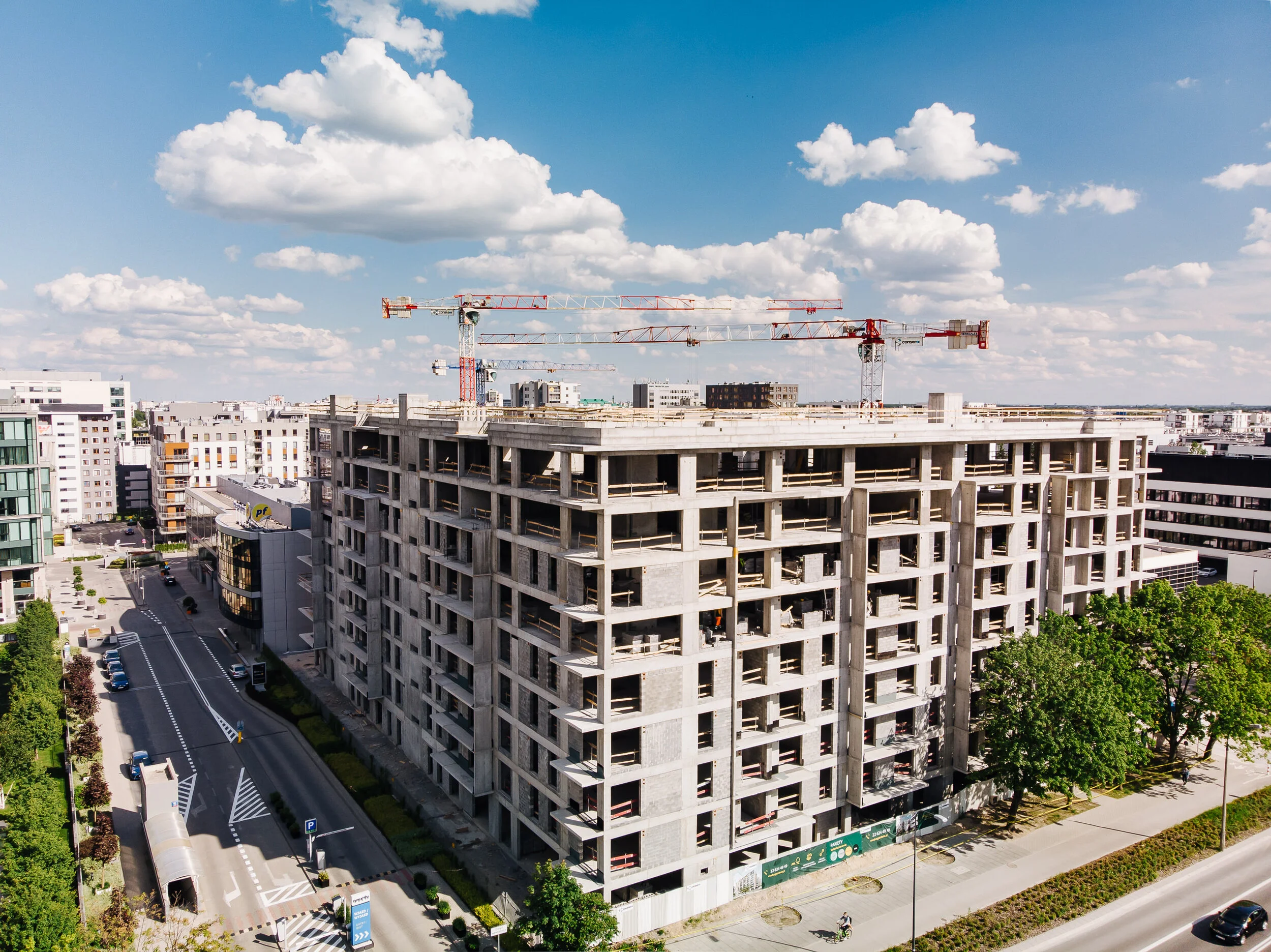 An unfinished multi-story building under construction in an urban area with two crane towers on top, surrounded by modern office and apartment buildings under a cloudy blue sky.