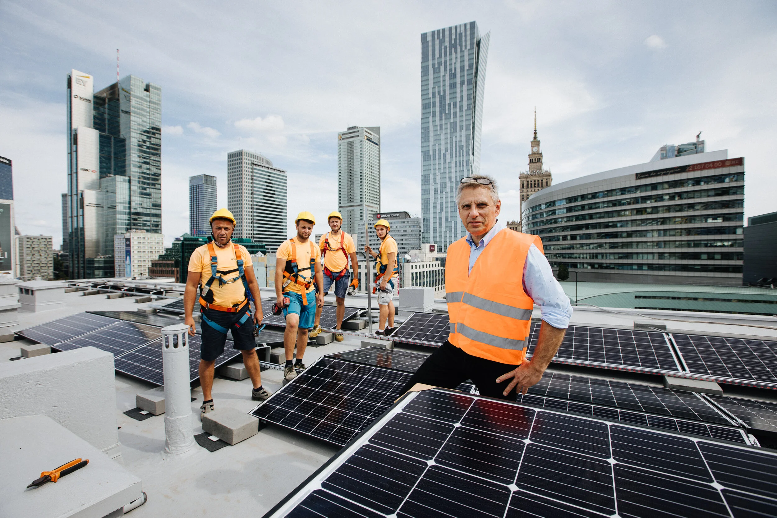 A group of five workers installing solar panels on a city rooftop with tall skyscrapers in the background. One man in the foreground is wearing an orange safety vest, and four other workers are wearing yellow helmets and safety harnesses.