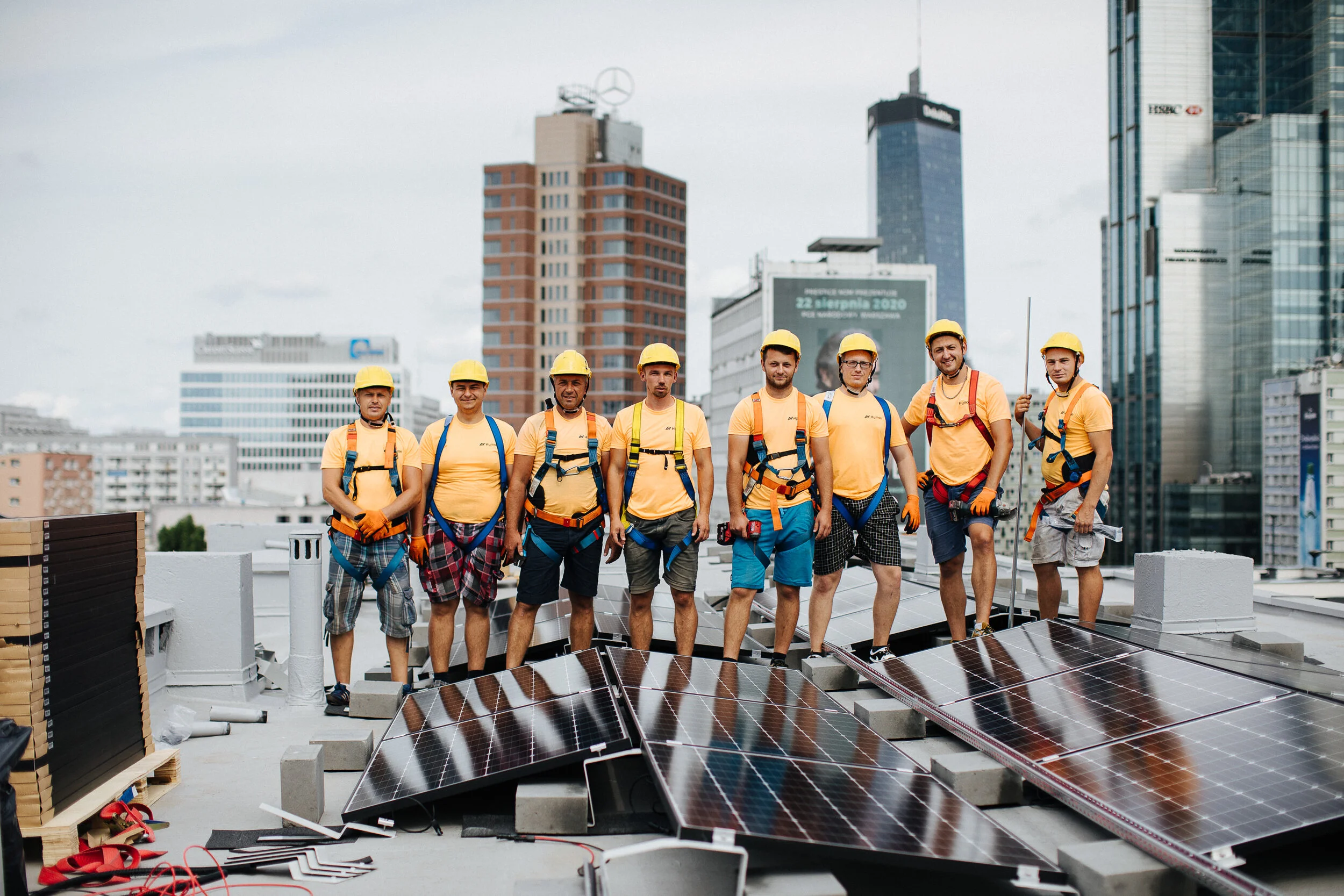 Group of construction workers standing on a rooftop with solar panels, wearing yellow helmets and safety harnesses, in an urban cityscape background.
