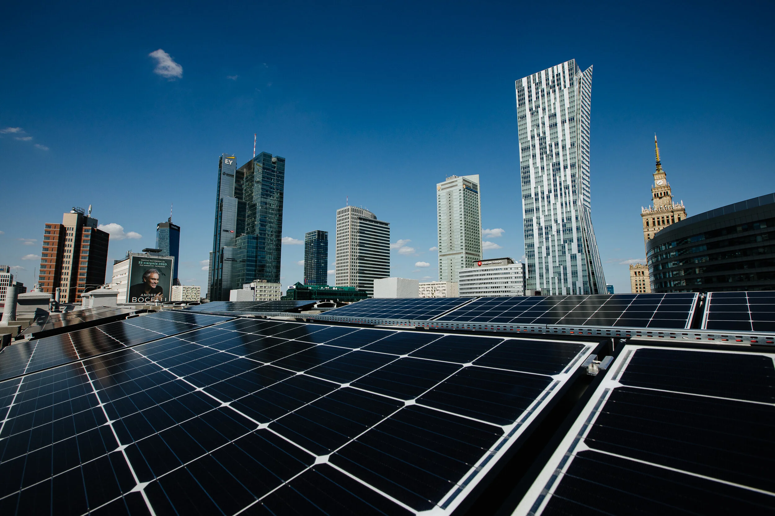 Solar panels on a rooftop with a city skyline in the background, featuring tall modern buildings under a clear blue sky.